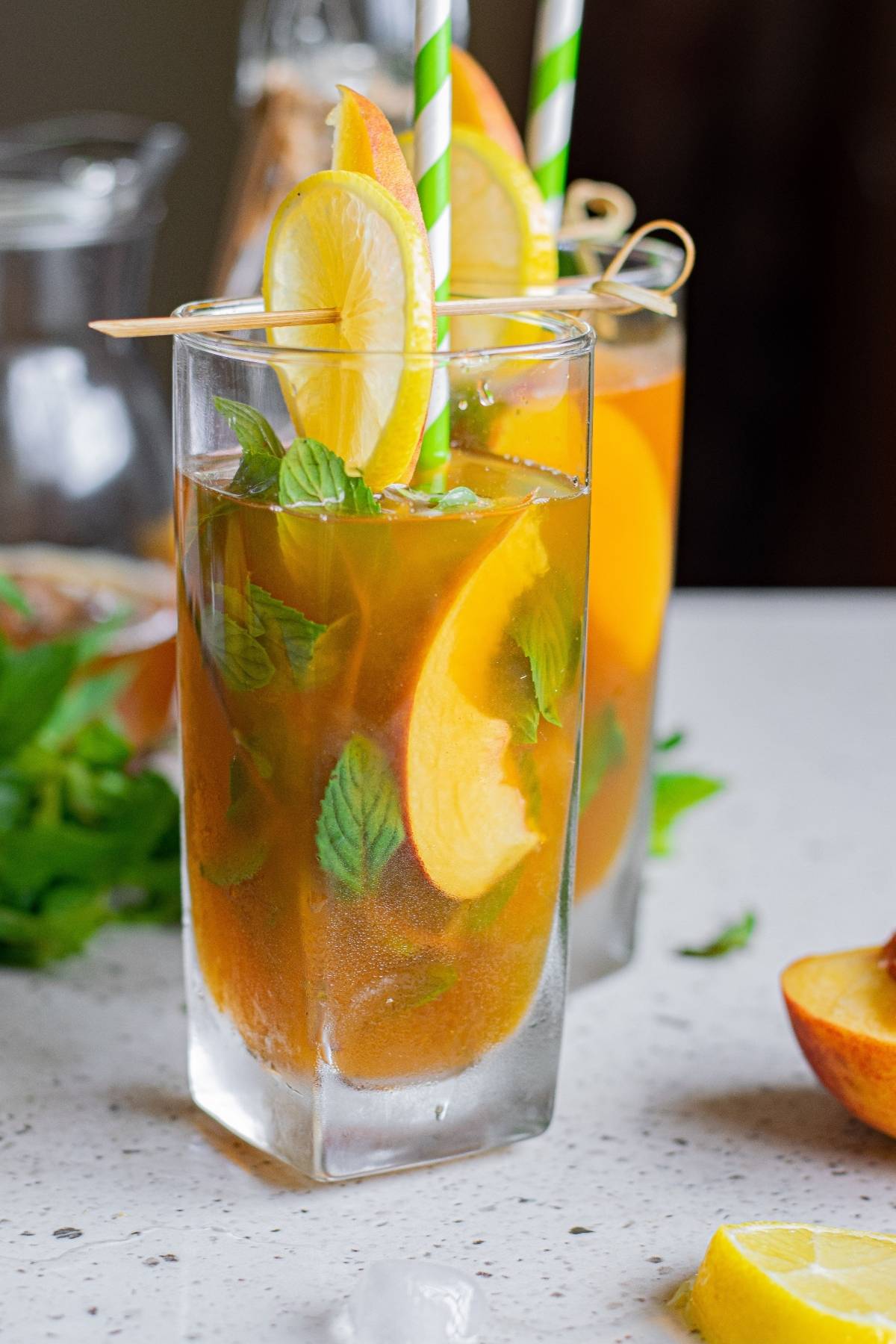 Iced tea with lemon and peach slices, mint leaves, and striped straws in tall glasses on a countertop—perfect for a refreshing alcoholic peach tea.