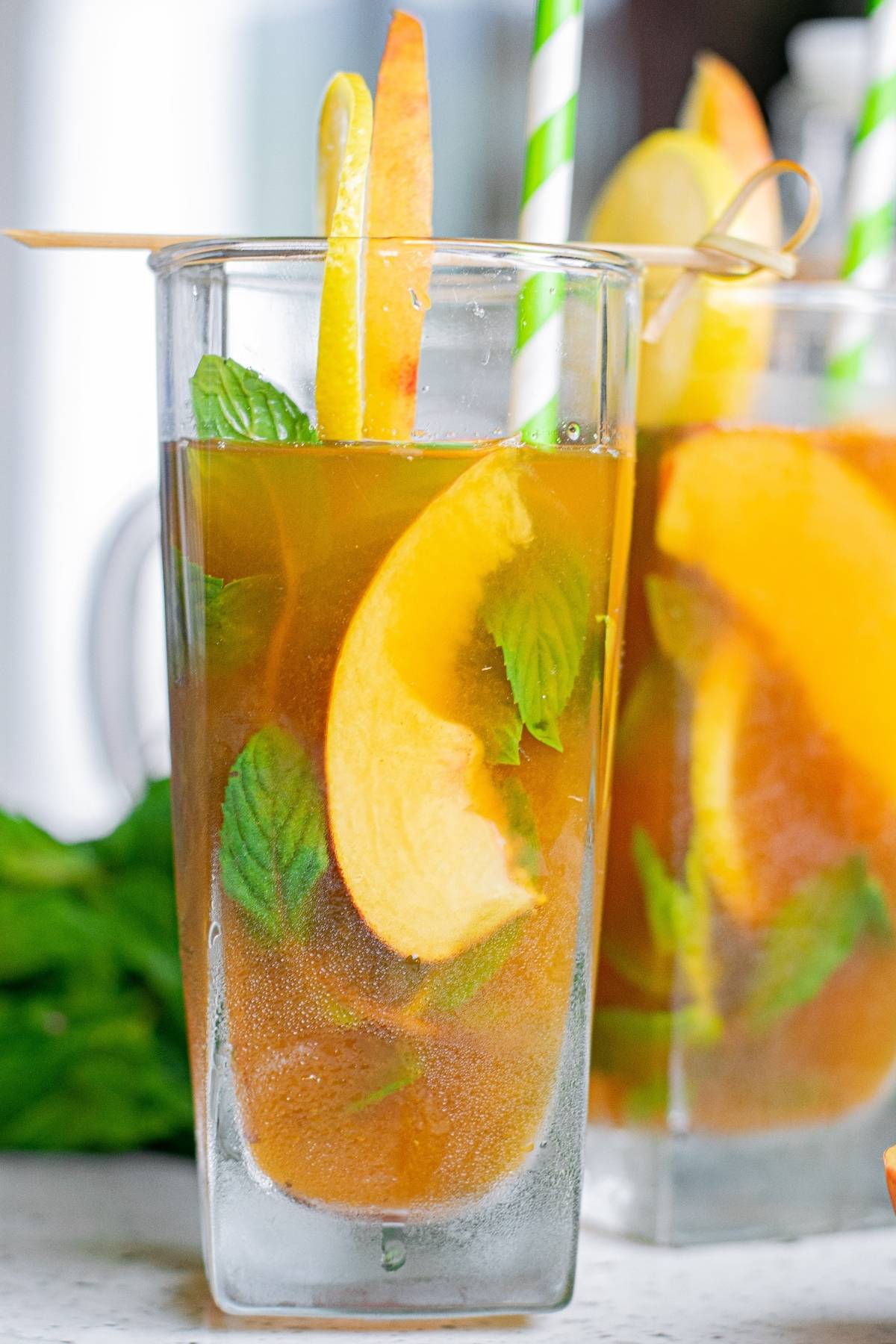 Iced tea and lemonade cocktail in a glass with peach slices, mint leaves, and green striped straws.