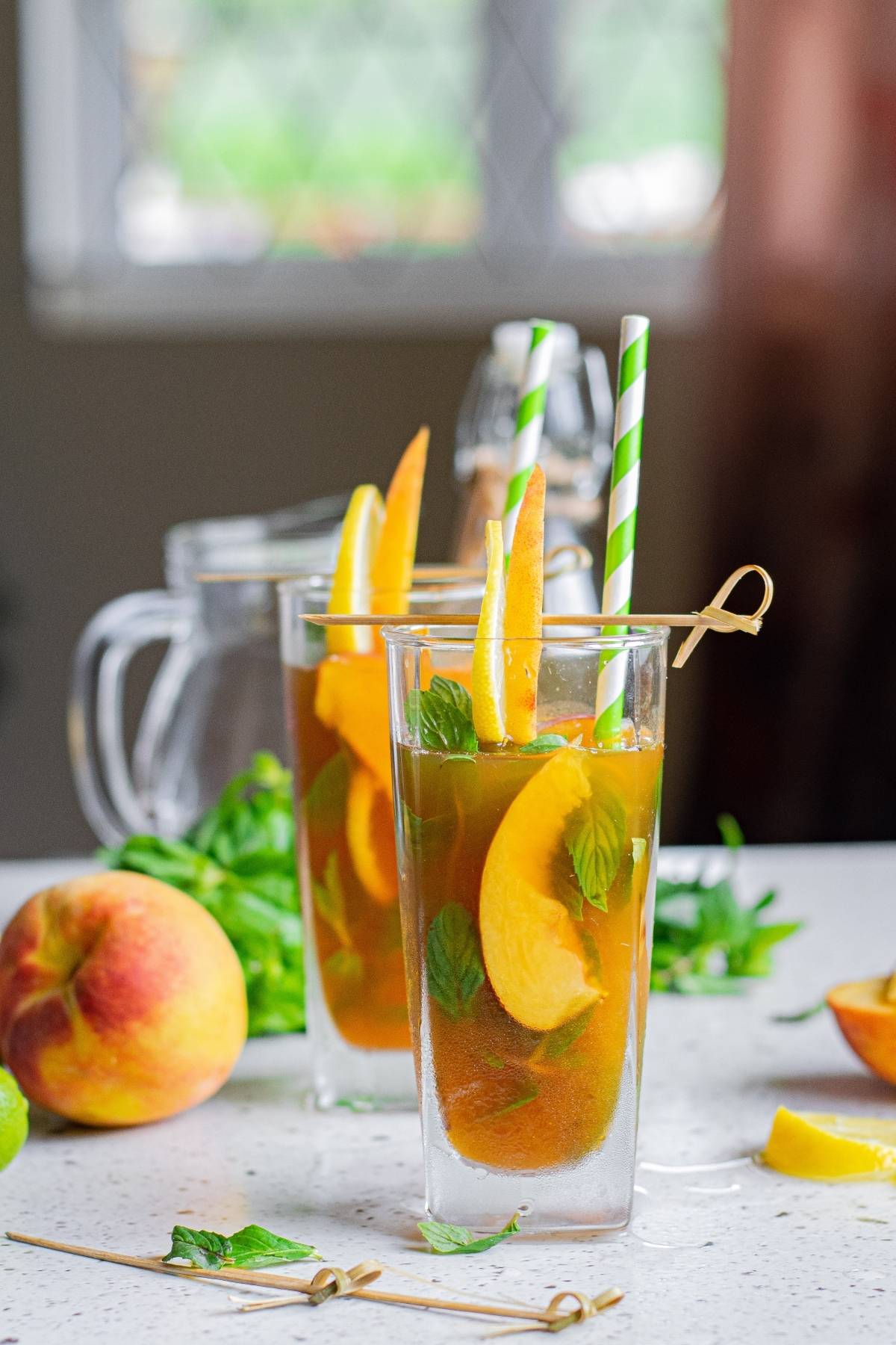 Two glasses of Hard Peach Iced Tea with peach slices, mint, and striped straws sit on a counter surrounded by fresh fruit.