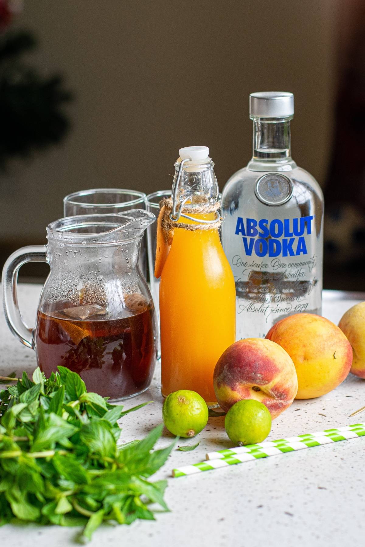 A bottle of vodka, iced tea, peach juice, peaches, limes, mint leaves, and straws on a kitchen counter—perfect for mixing a refreshing boozy iced tea recipe.