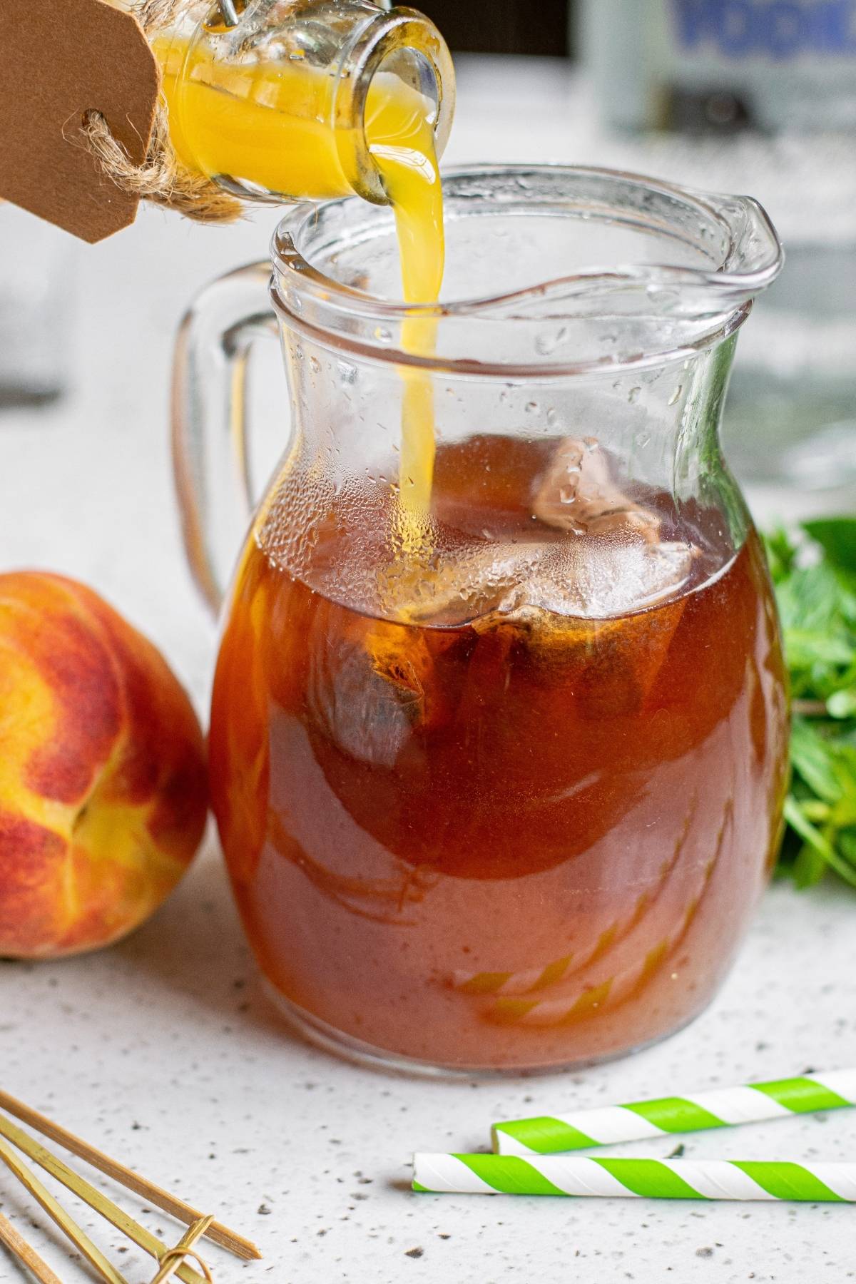 Peach juice is poured into a glass pitcher of boozy iced tea, with a fresh peach and green-striped straws nearby—a refreshing alcoholic peach tea perfect for summer sipping.