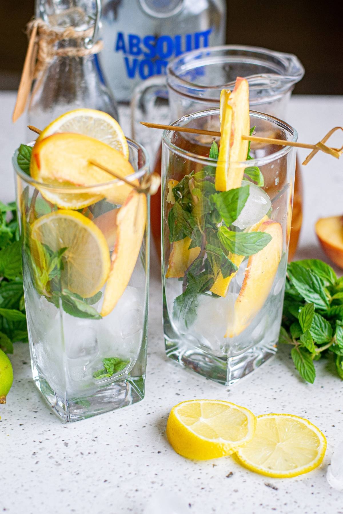 Two tall glasses with ice, lemon, peach slices, and mint showcase a refreshing hard spiked iced tea lemonade, with vodka and fresh lemon on the counter.
