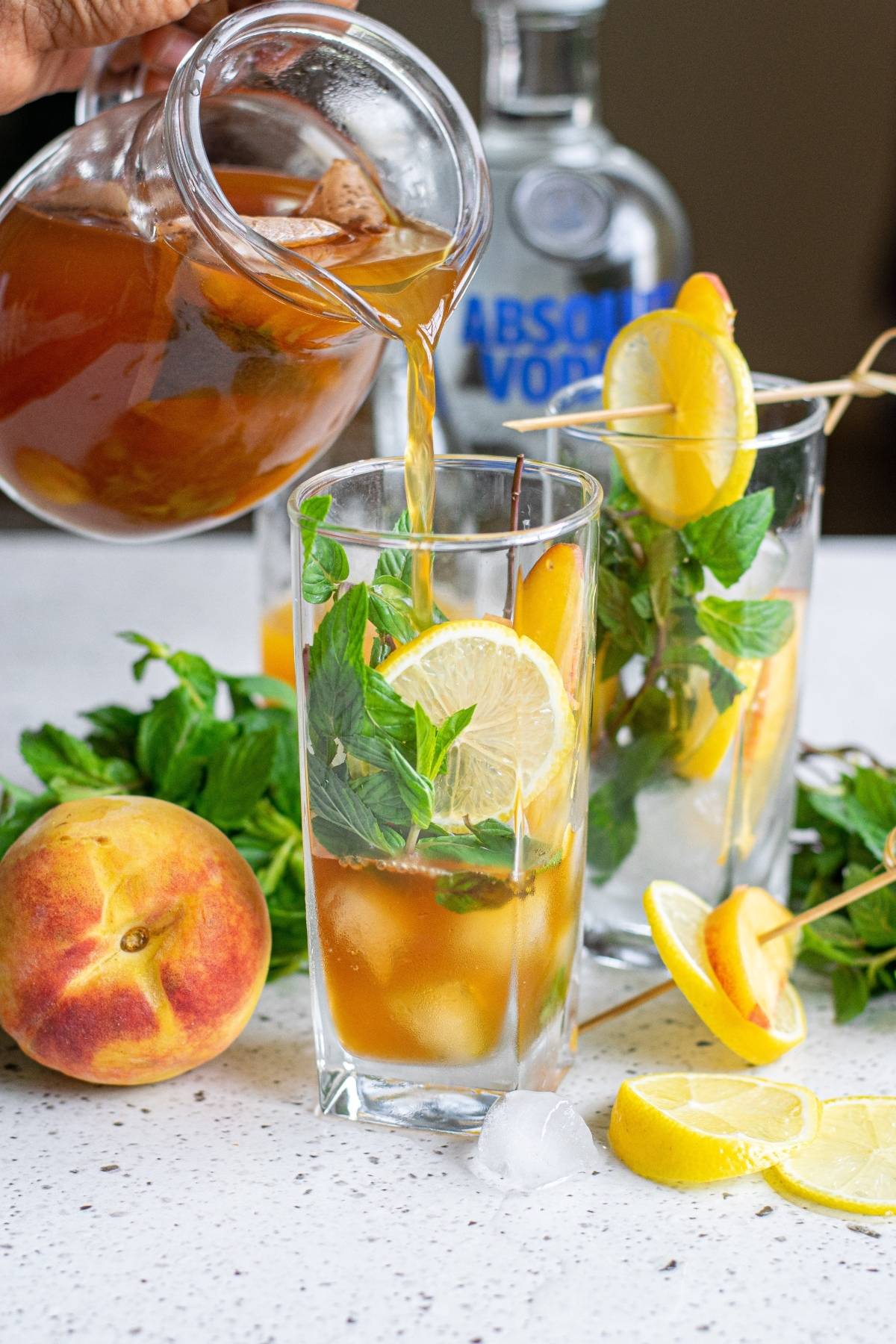 Hard Peach Iced Tea with lemon, mint, and peach is poured into a glass next to vodka, fresh peaches, and lemons.