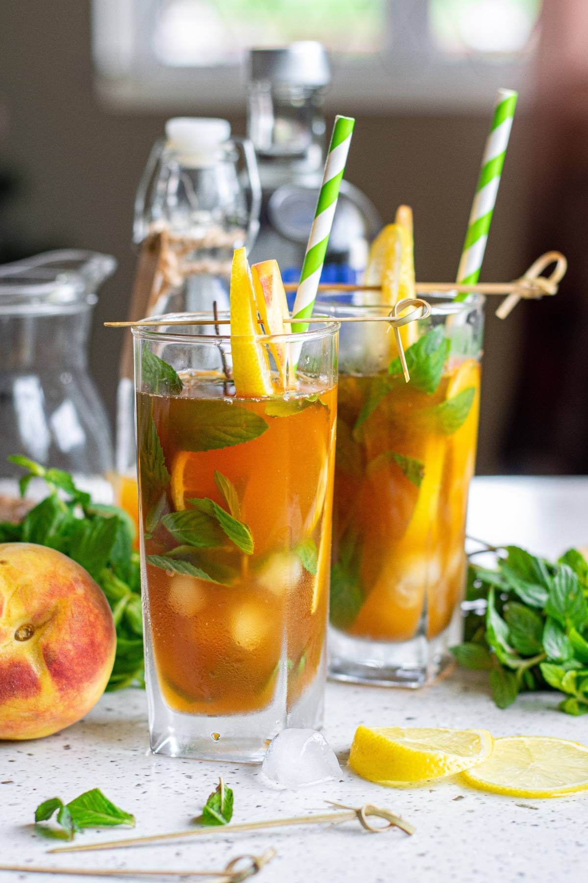Two glasses of iced tea and lemonade cocktail with lemon, mint, and striped straws, surrounded by peach, mint, and lemon slices.
