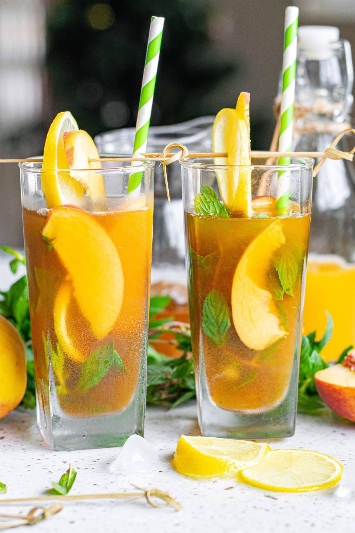 Two glasses of Hard Peach Iced Tea with lemon, peach slices, mint, and striped straws sit on a light surface.