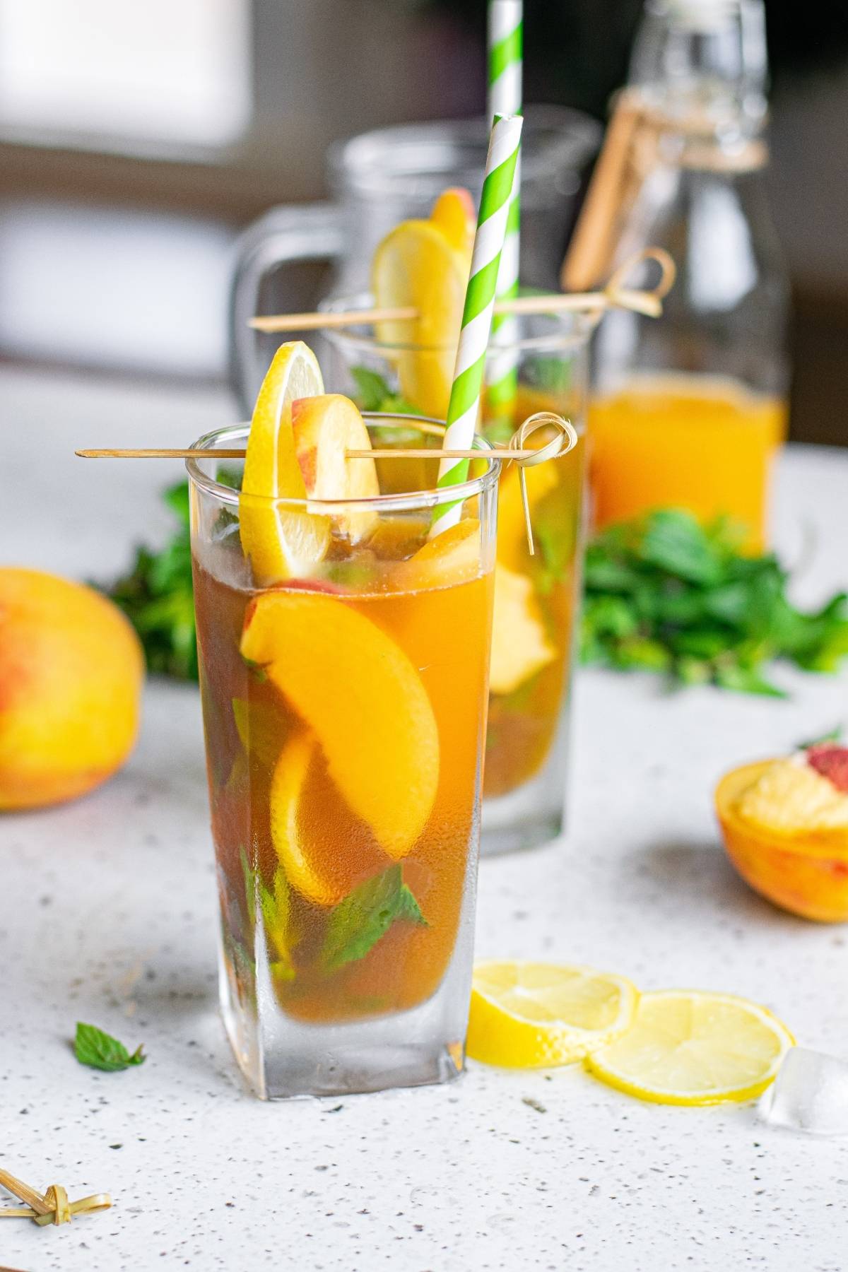 Two tall glasses of Hard Peach Iced Tea with peach slices, lemon, mint, and striped straws sit on a white countertop.