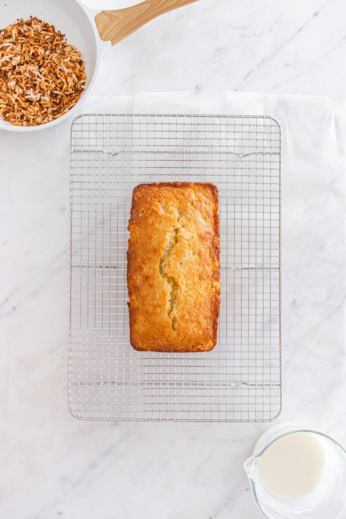 A loaf of bread cools on a wire rack, with a bowl of toasted coconut and a glass of milk nearby.