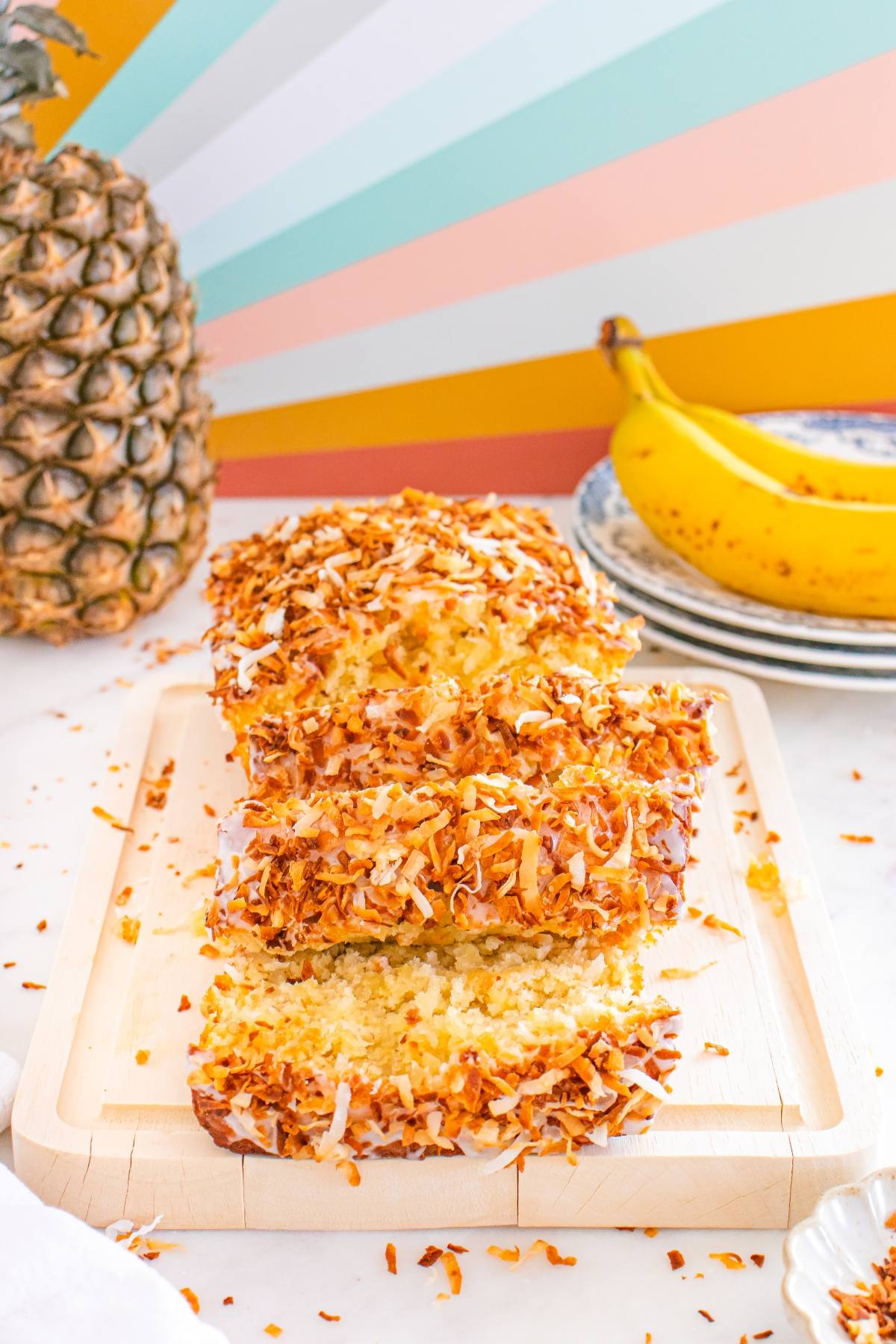 A sliced loaf of coconut-topped banana bread on a wooden tray, with a pineapple and banana in the background.