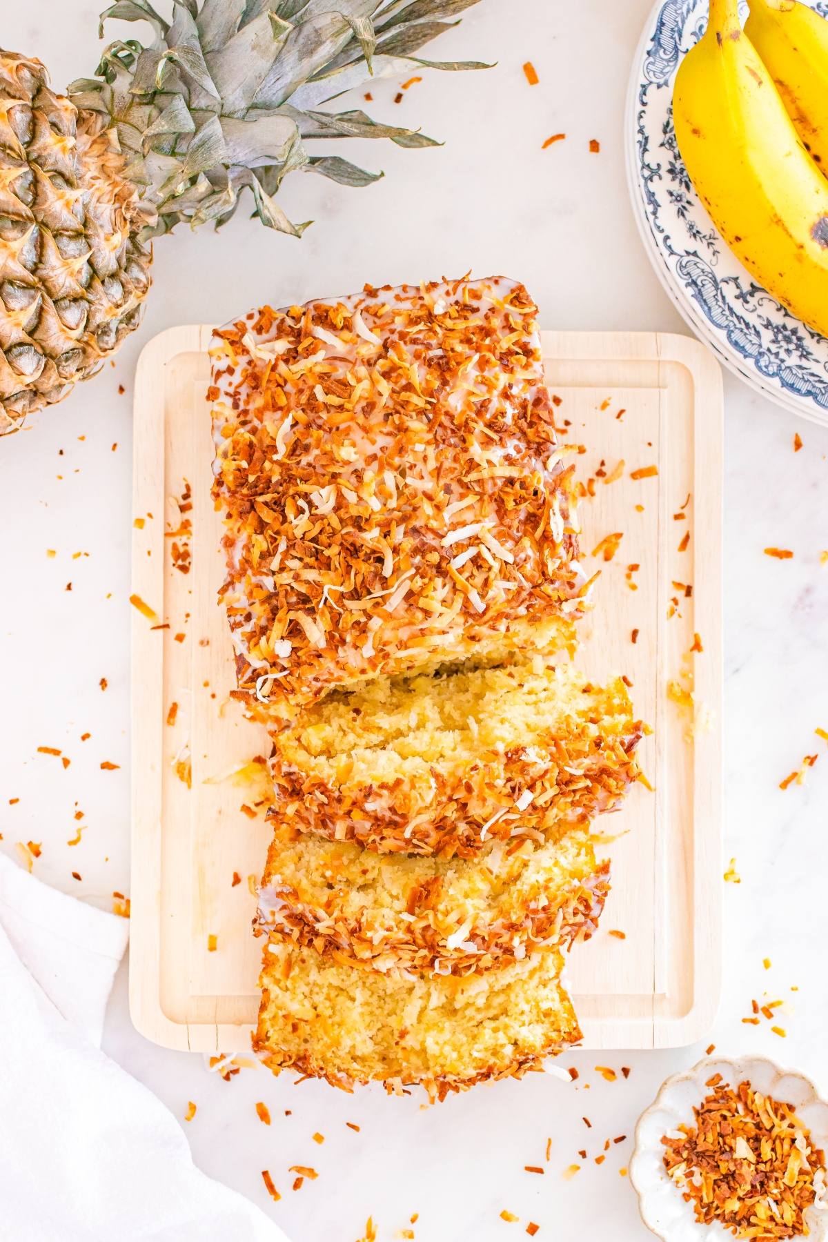 Sliced coconut bread with toasted coconut topping on a cutting board, surrounded by pineapple and bananas.