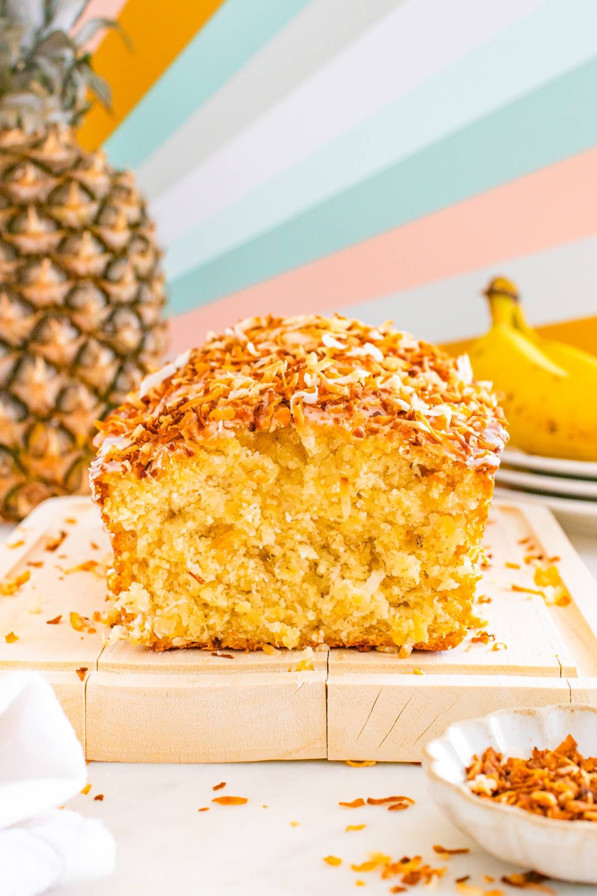A loaf of coconut bread with a golden crust, sliced to show its moist texture, with pineapple and bananas behind.