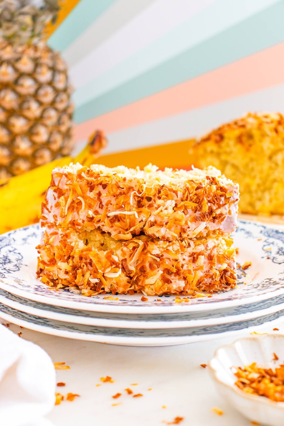 Two slices of toasted coconut cake on a plate, with a pineapple and banana in the background.