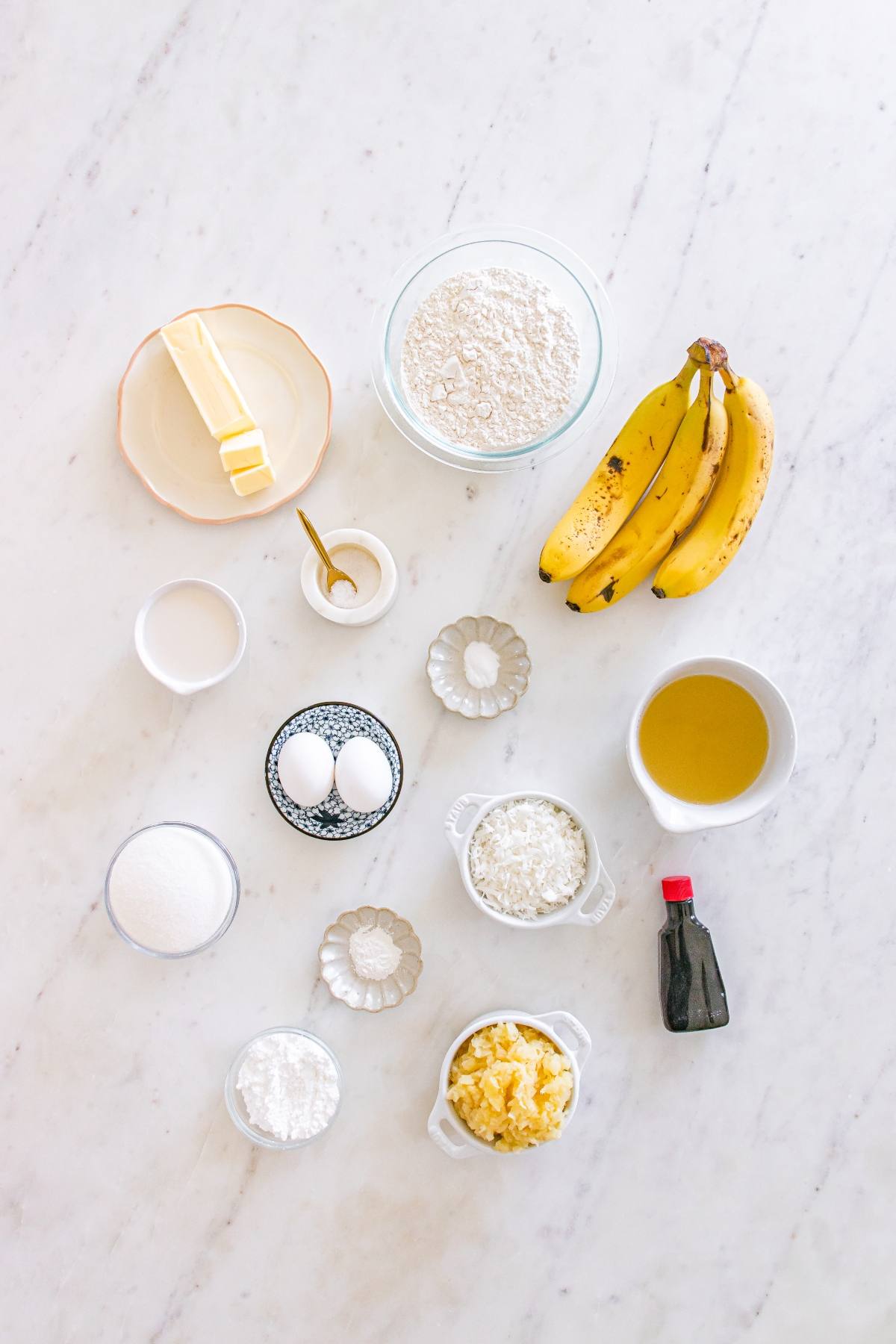 Baking ingredients on a white surface: bananas, flour, butter, eggs, oil, sugar, and small bowls with other items.