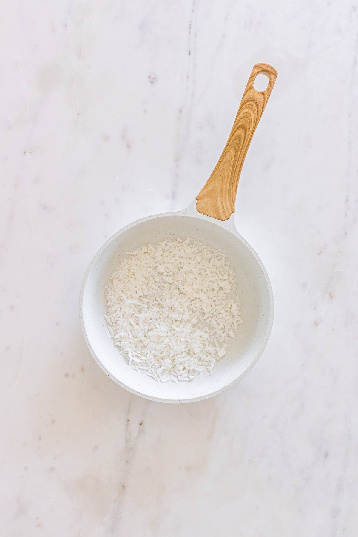 A white pan with a wooden handle containing shredded coconut on a white marble surface.