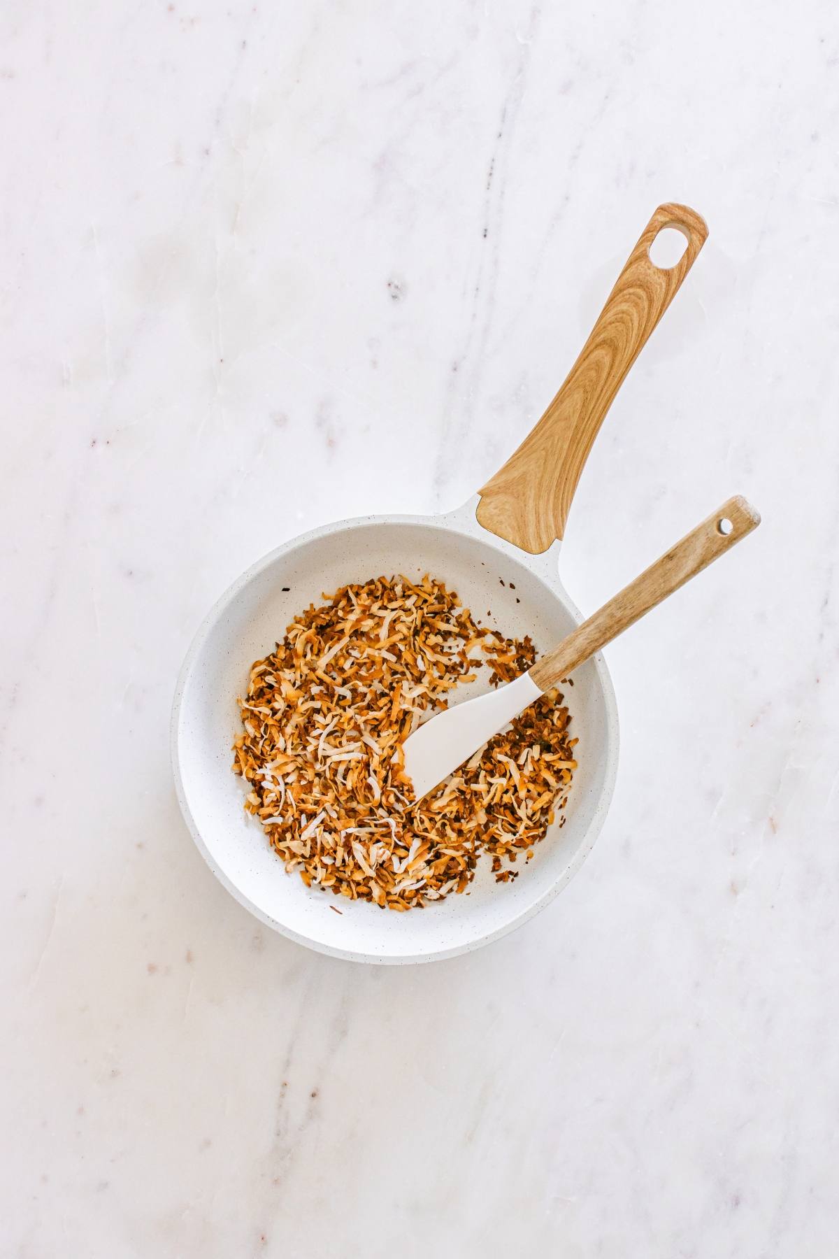 A white pan with toasted shredded coconut and a spatula on a white marble surface.