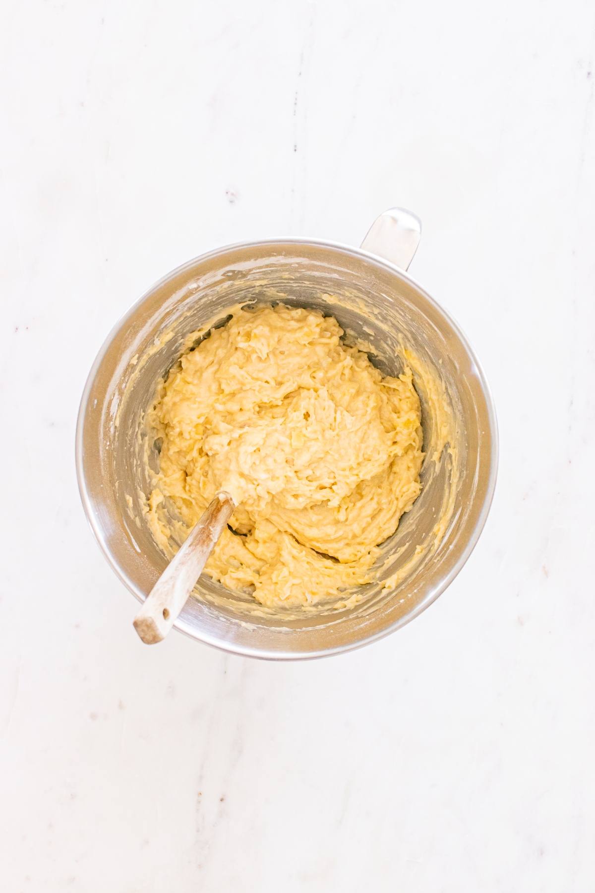 A metal mixing bowl filled with thick, yellow batter and a wooden spoon on a white surface.