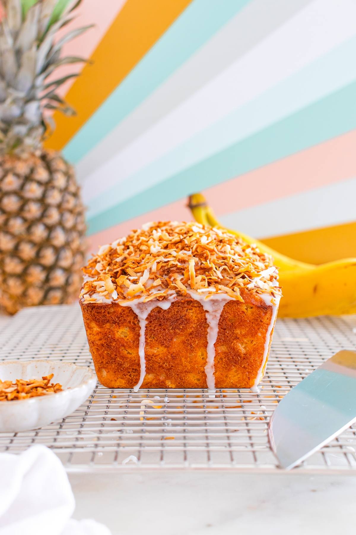 A glazed loaf topped with toasted coconut sits on a cooling rack, with pineapple and bananas in the background.