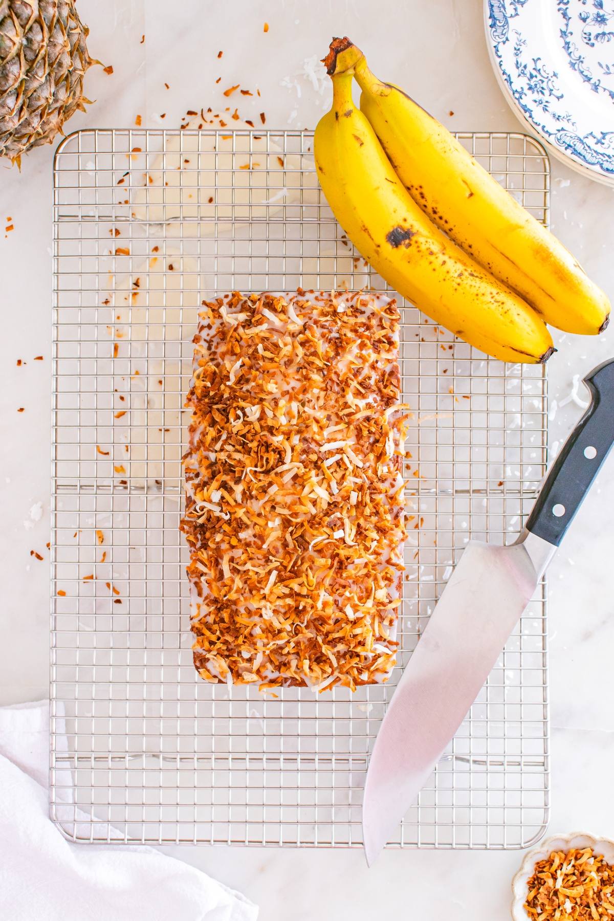 A loaf topped with toasted coconut sits on a cooling rack beside ripe bananas and a large knife.