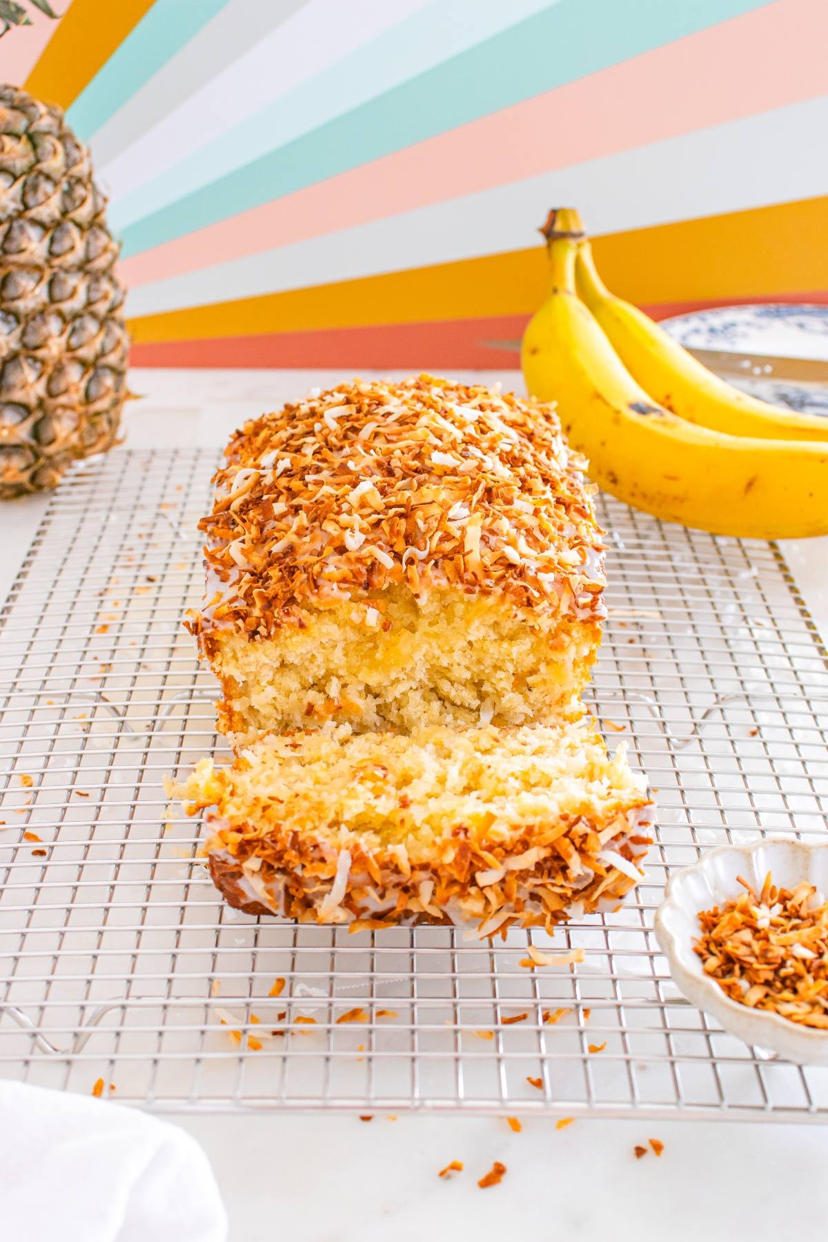 A sliced loaf of toasted coconut banana bread on a cooling rack with bananas and a pineapple in the background.