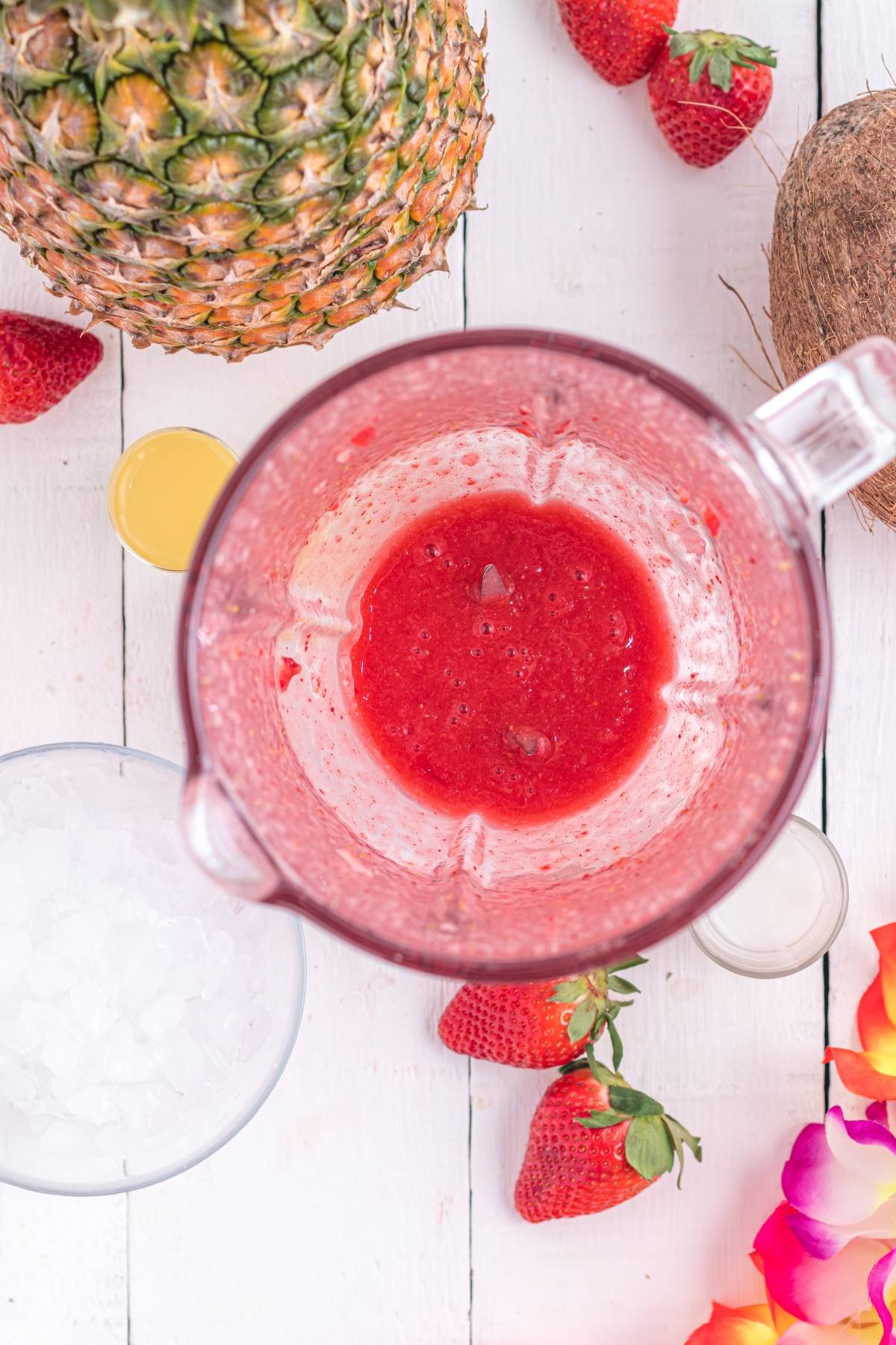A blender with strawberry puree, surrounded by strawberries, pineapple, coconut, ice, and a flower—perfect for mixing a Hawaiian Lava Flow Cocktail or your favorite Hawaiian tropical drink.