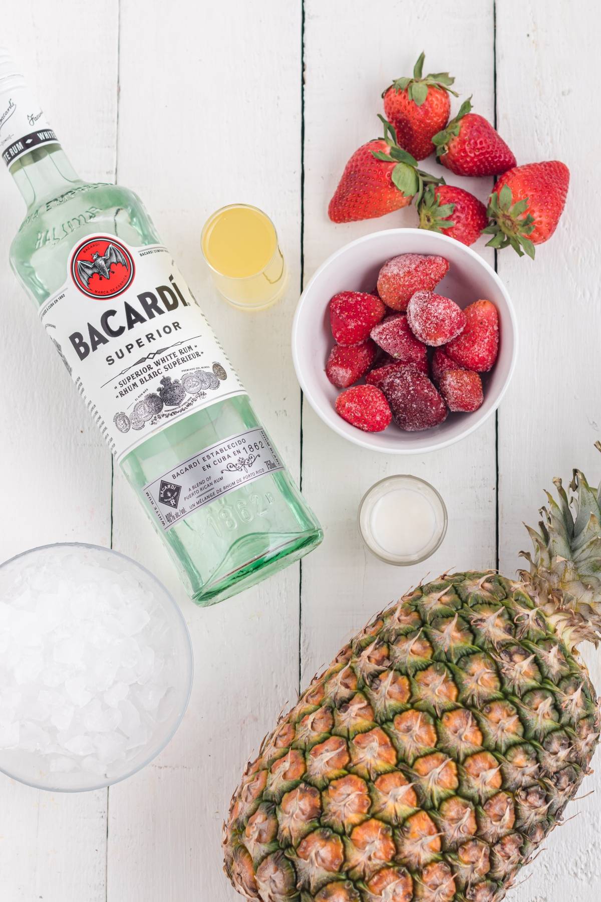 Bacardi rum, fresh and frozen strawberries, pineapple, ice, and shot glasses on a white wooden table—perfect for mixing a Hawaiian Lava Flow Cocktail or another tropical cocktail with cream of coconut.