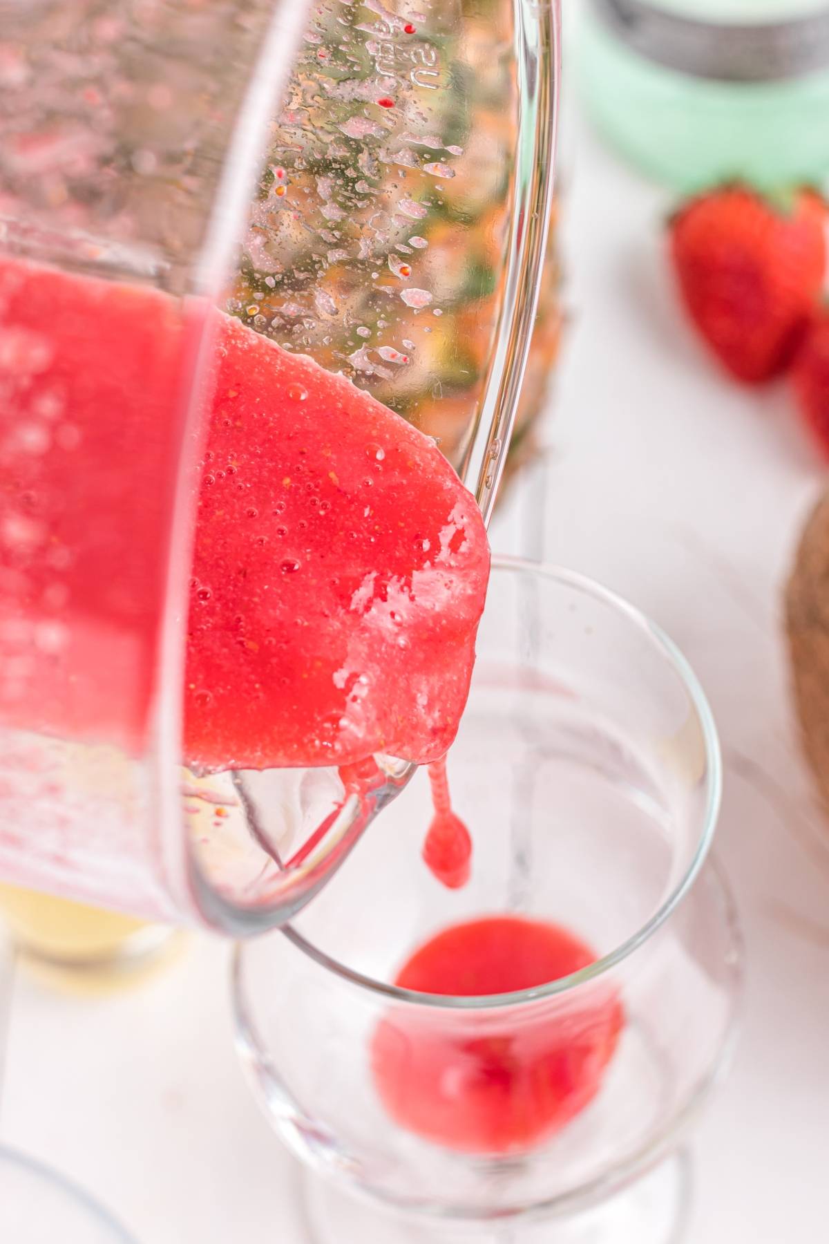 A vibrant Hawaiian Lava Flow Cocktail, with bright red strawberry smoothie and cream of coconut, is poured from a blender into a clear glass, surrounded by fresh strawberries.