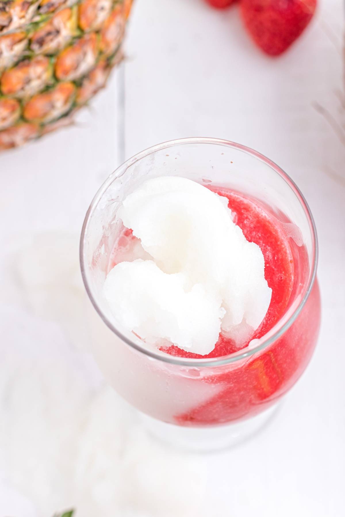 A glass of Hawaiian lava flow—a cocktail with cream of coconut, white and red slush—sits beside a pineapple and strawberries on a white surface.