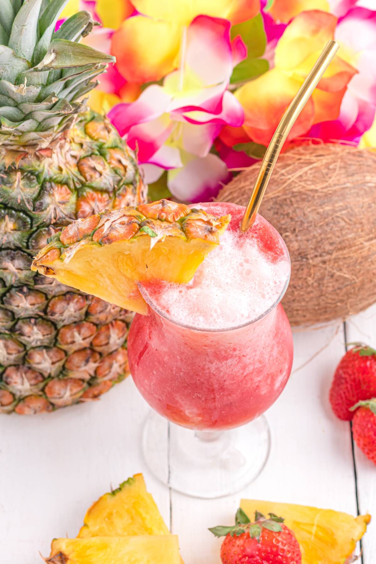 A Hawaiian lava flow cocktail with cream of coconut, garnished with a pineapple slice and surrounded by fresh pineapple, coconut, strawberries, and colorful flowers.