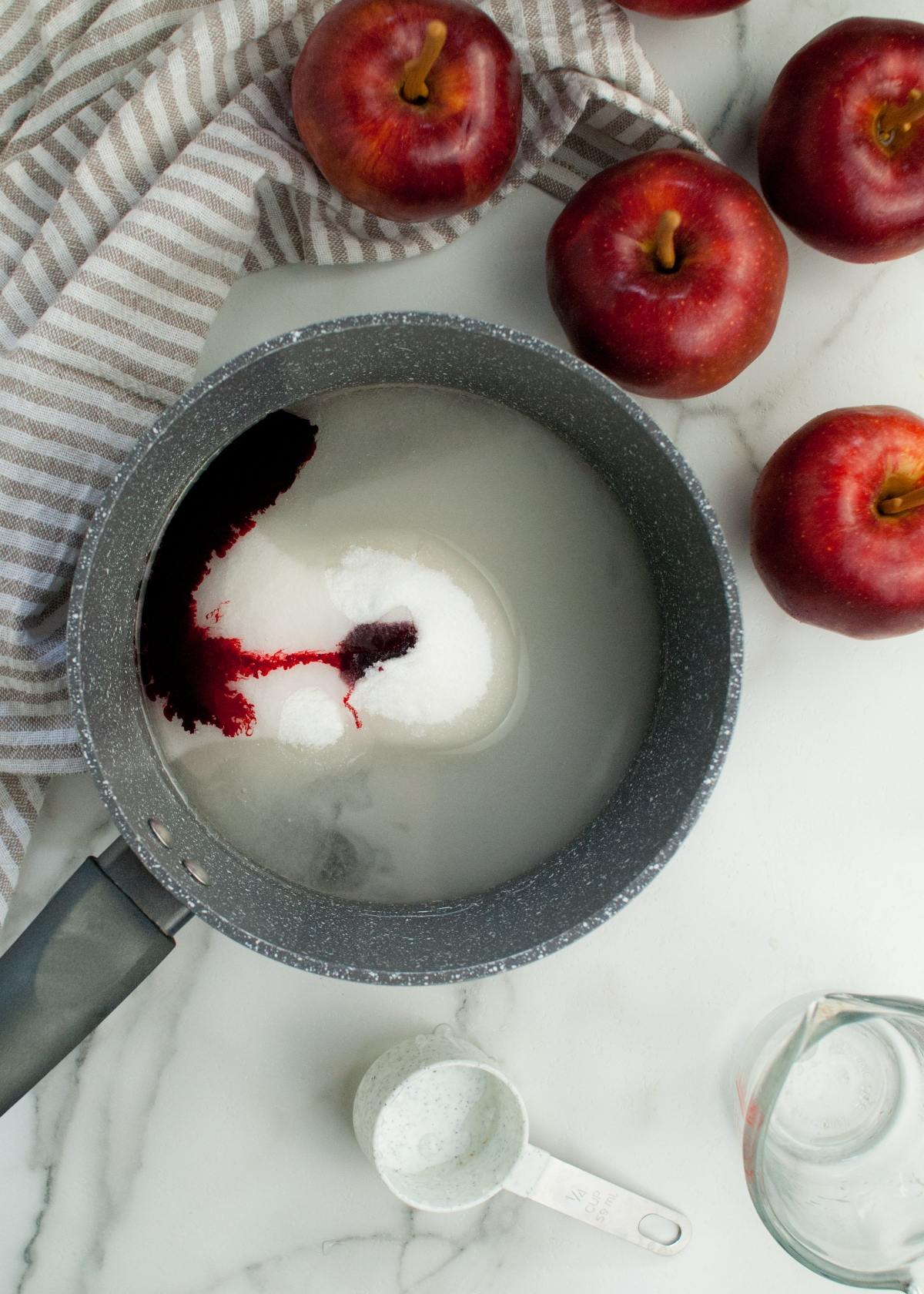 A saucepan with sugar and red liquid, surrounded by red apples on a marble surface with a striped towel—a perfect setup for making the best candied apples or learning how to make candy apples on a stick.
