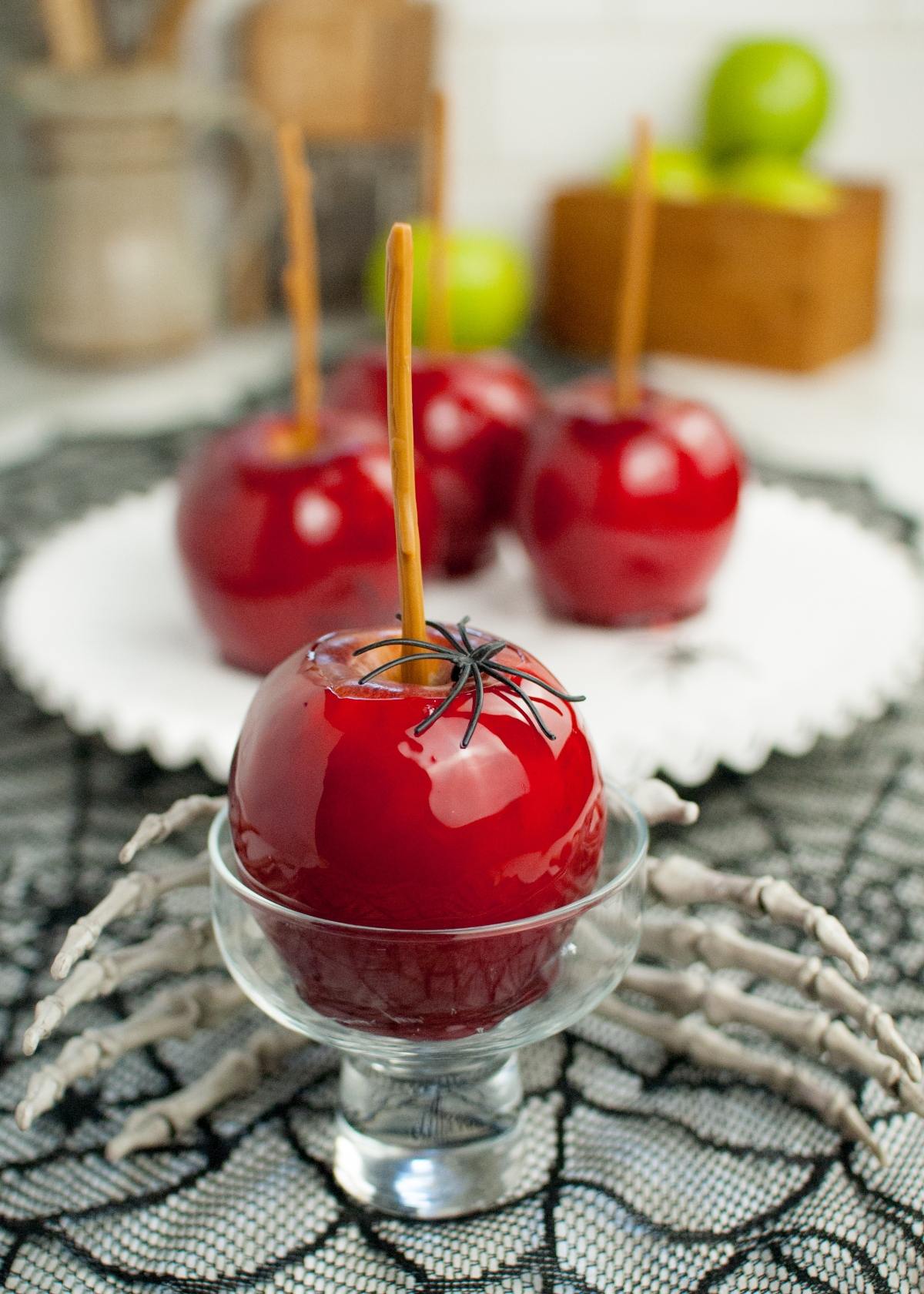 Glossy red candy coated apples with a plastic spider on top, displayed in a glass dish with a skeleton hand prop, make for the best candied apples at any spooky party.