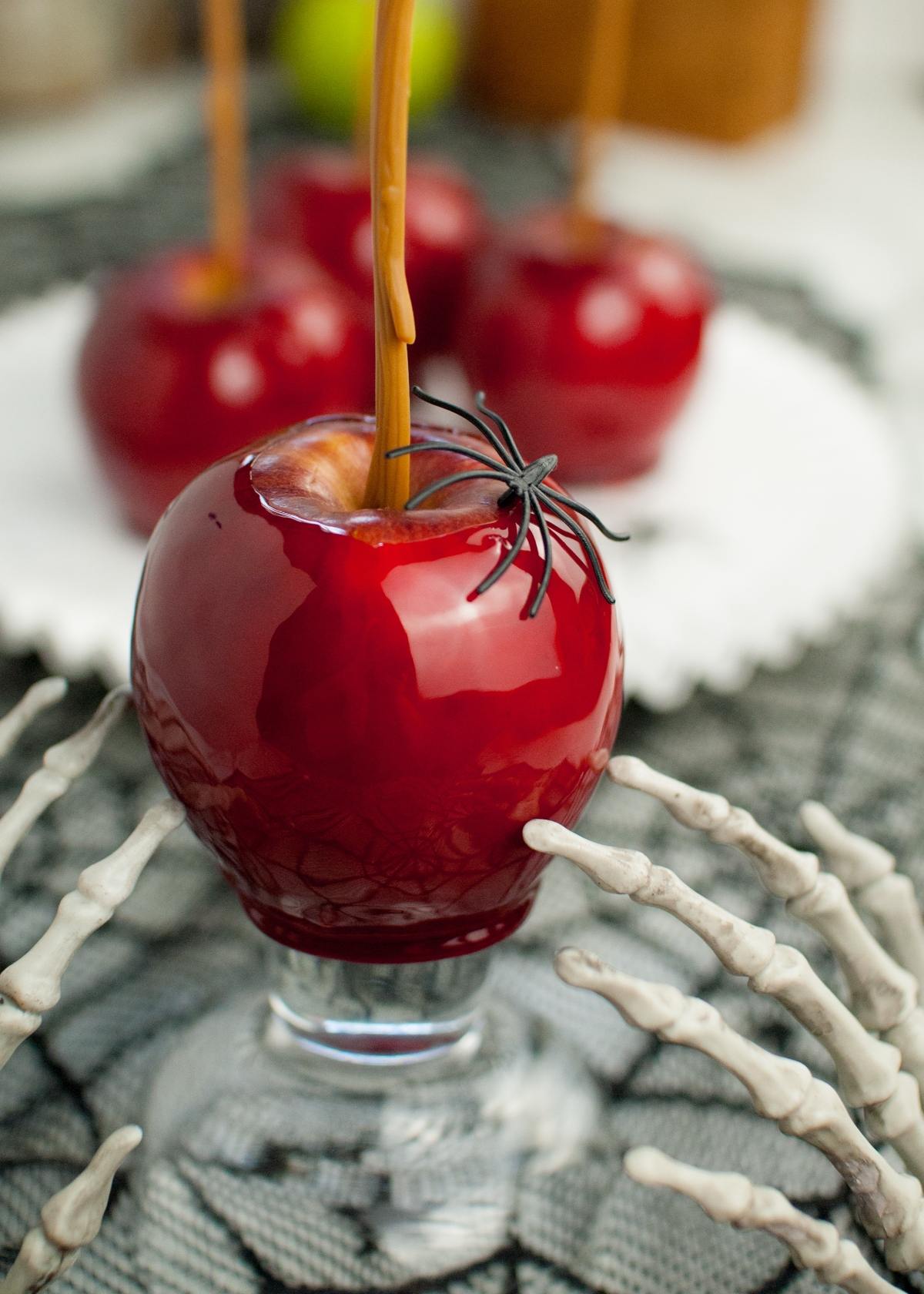 A shiny red candy coated apple with a fake spider and skeleton hands, with more apples in the background—perfect for anyone craving the best candied apples at home.