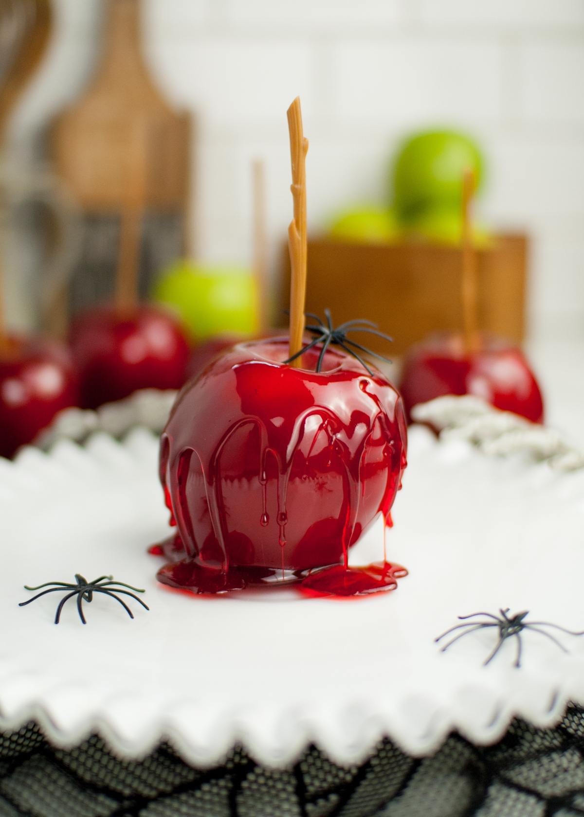 A glossy red candy coated apple with a fake black spider sits on a white plate, with more homemade candy apples and spiders in the background.
