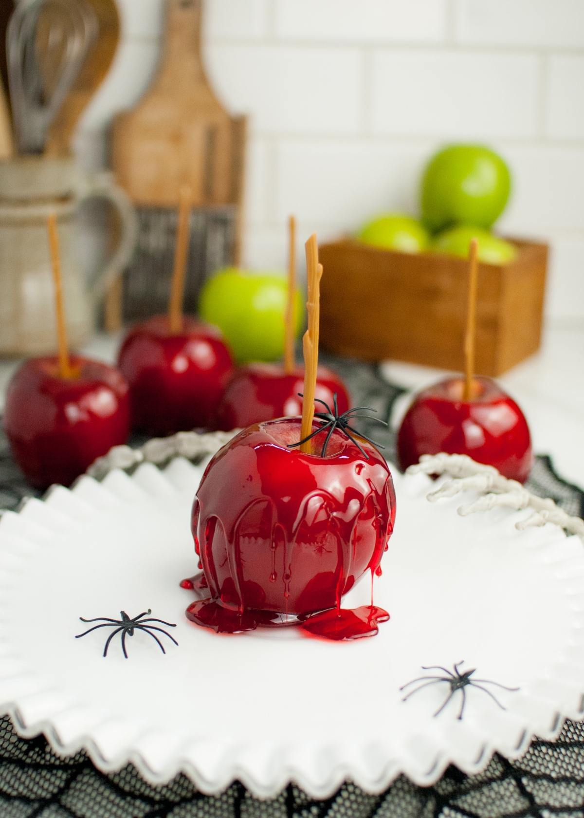 A shiny red candy coated apple on a white plate with toy spiders, with more homemade candy apples and green apples in the background.