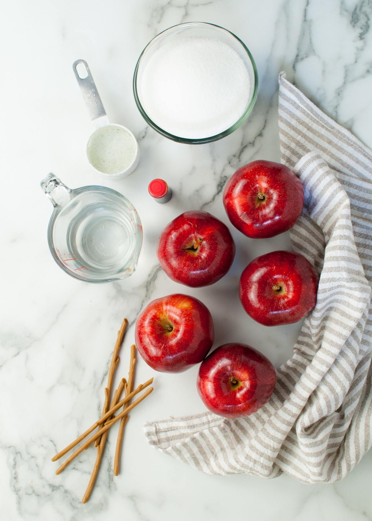Five red apples, a bowl of sugar, water, sticks, and a striped towel on a marble surface—everything you need for homemade candy apples or learning how to make candy apples on a stick.