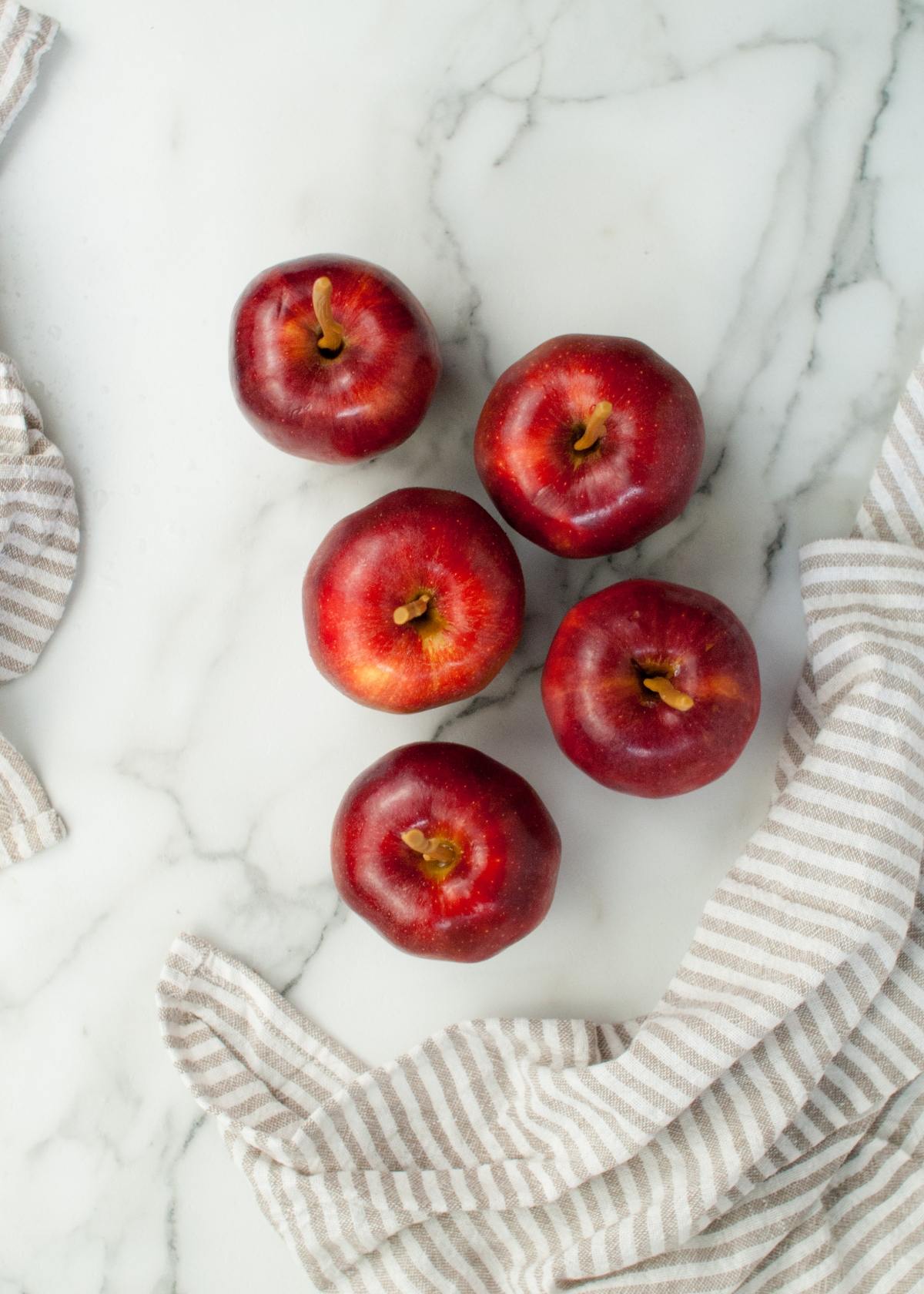 Five red candy coated apples rest on a marble surface, with two beige-striped cloths at the corners, capturing the classic look of the best candied apples.
