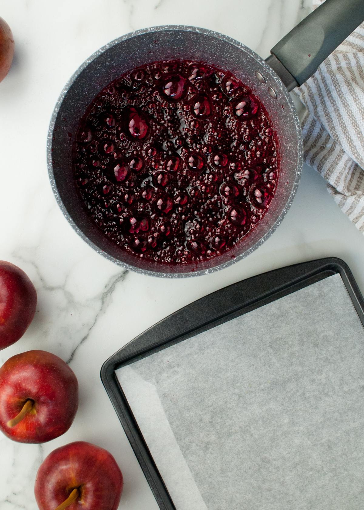 A saucepan of simmering berry sauce sits beside plums and a parchment-lined baking sheet, ready to craft the best candied apples or delicious red candy coated apples on a marble surface.