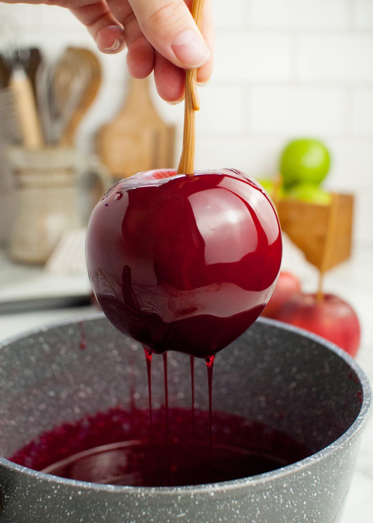 A hand dips an apple on a stick into shiny red candy coating over a pot in a kitchen, creating the best candied apples.