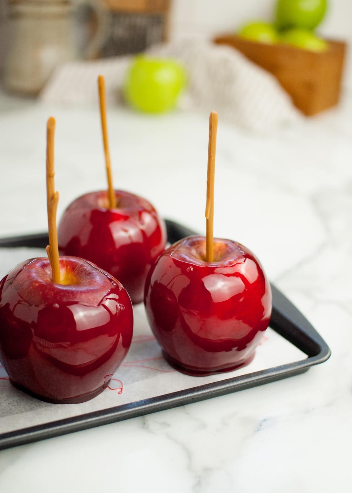 Three shiny red homemade candy apples with sticks on a tray, set on a white marble surface—perfect for anyone wanting the best candied apples at home.