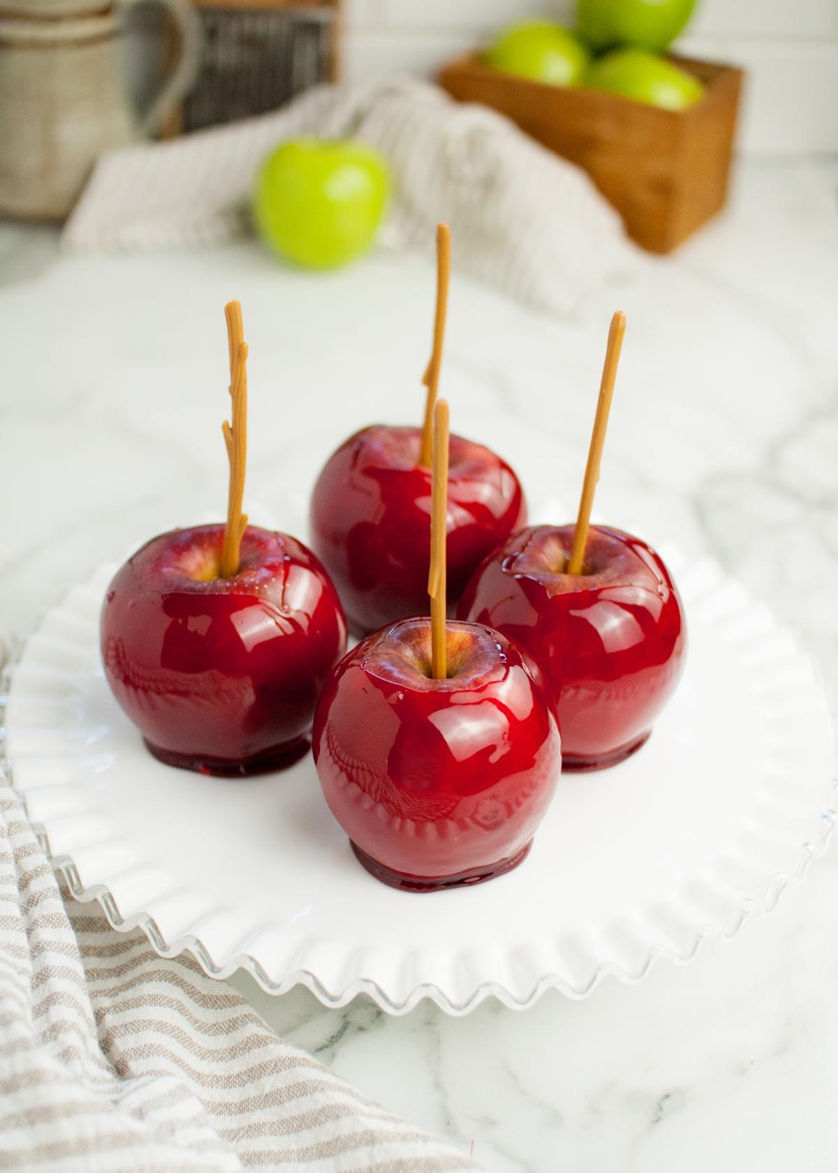 Four shiny red candy coated apples with sticks rest on a white plate, with green apples in the background—a perfect showcase for homemade candy apples.