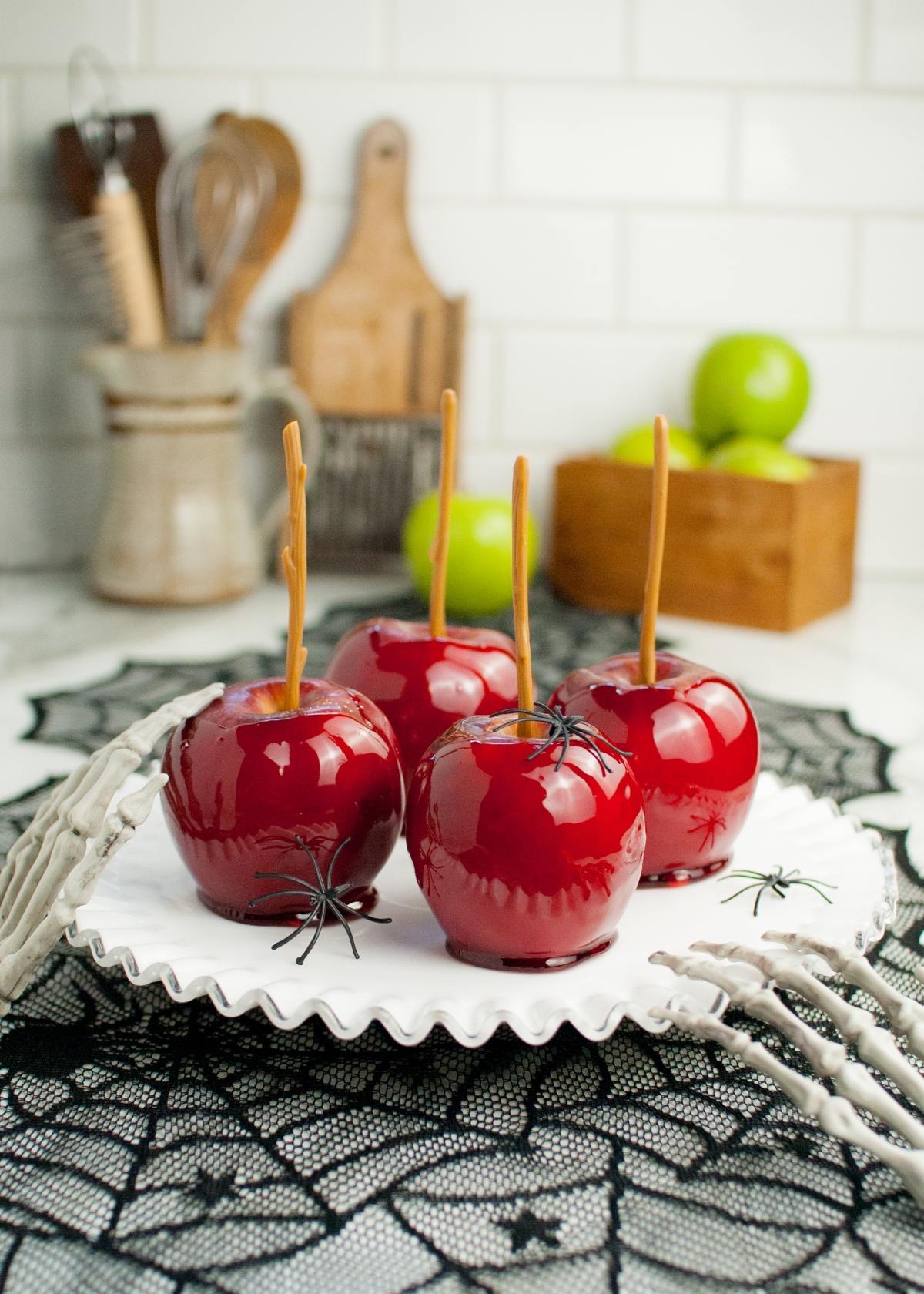 Four shiny red homemade candy apples with fake spiders on a white plate, set on a black lacy spiderweb cloth.