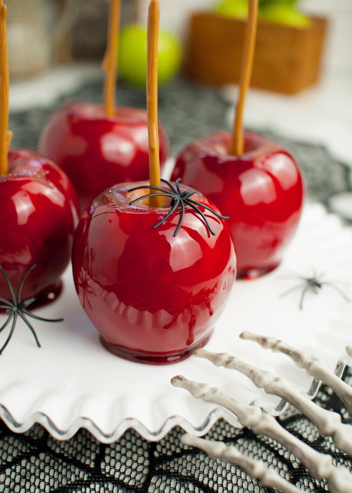 Shiny red homemade candy apples with fake spiders on top, displayed on a white plate with a skeleton hand nearby.