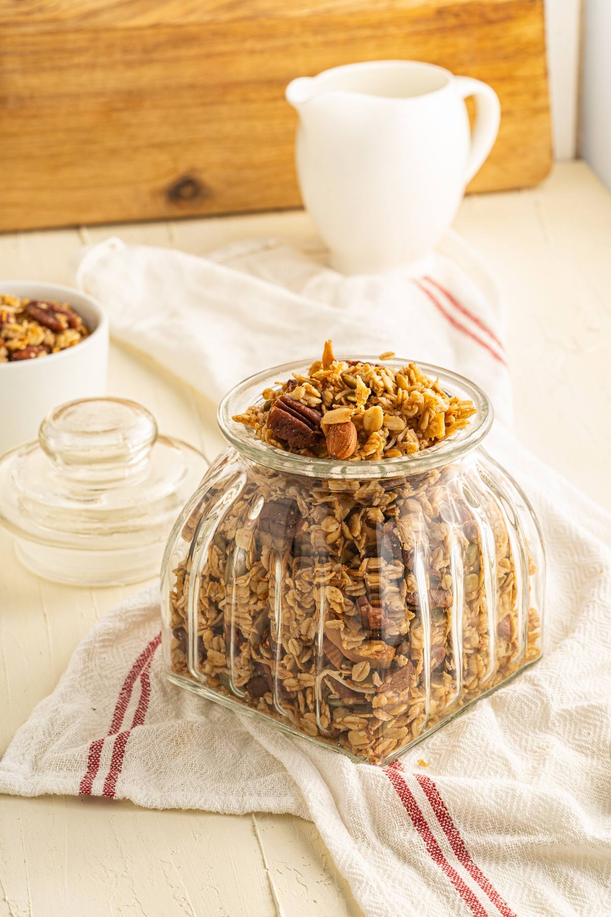 A glass jar of easy homemade granola sits on a white towel, with a small bowl and a white pitcher in the background—perfect as part of a healthy breakfast.