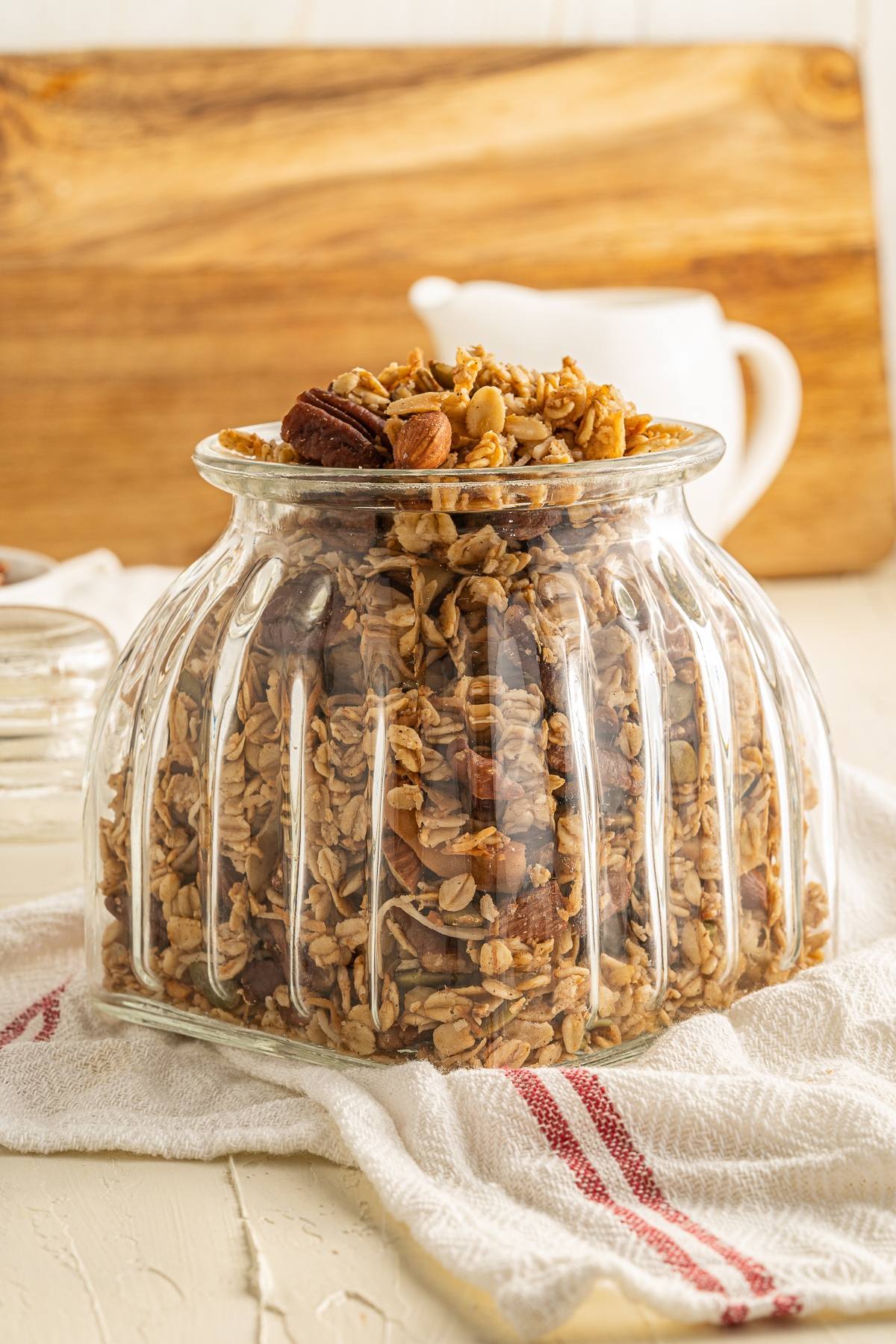 A glass jar filled with easy homemade granola sits on a white towel, with a wooden board and pitcher in the background.