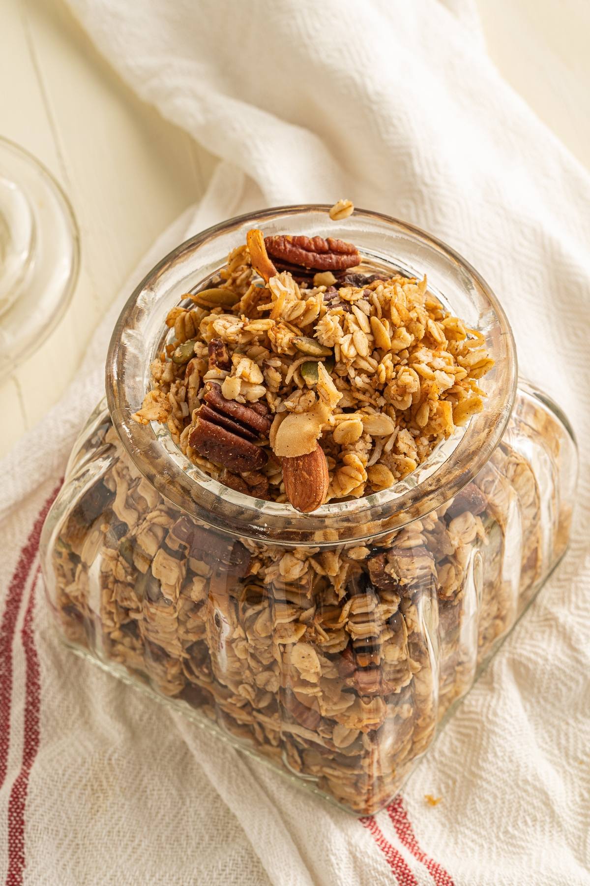 A glass jar filled with easy homemade granola, nuts, and seeds sits on a white towel with red stripes.