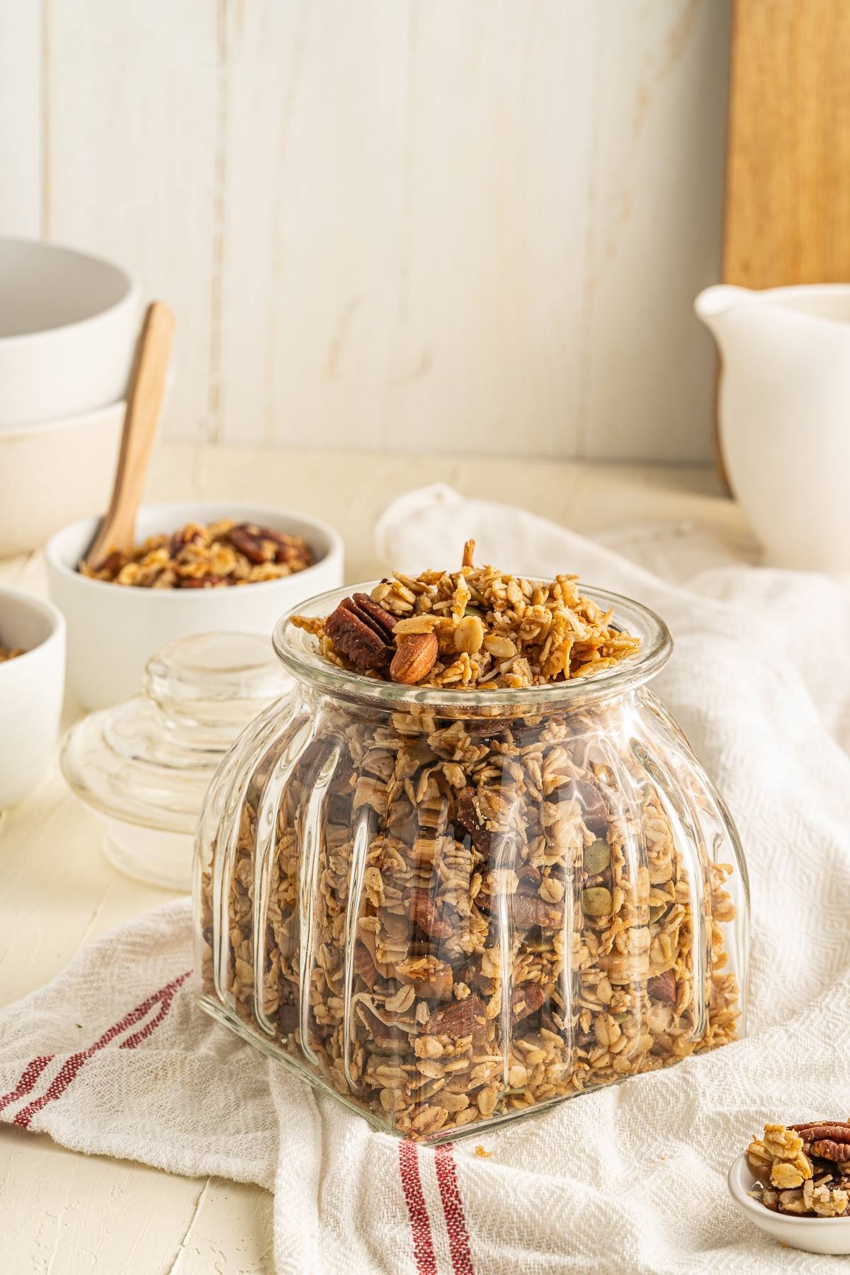 A glass jar filled with easy homemade granola sits on a white towel, with bowls and a pitcher in the background—part of a healthy breakfast.