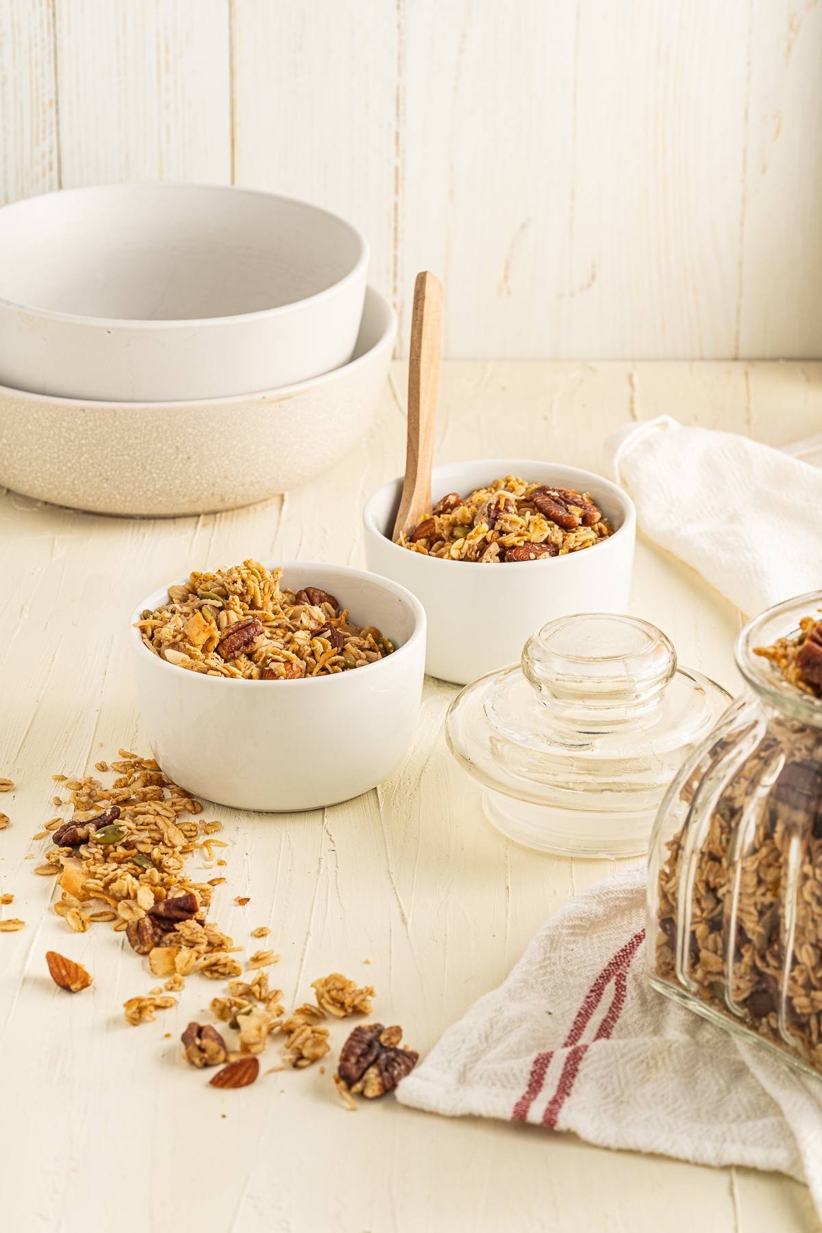 Two bowls of easy homemade granola with nuts and a wooden spoon, surrounded by stacked bowls and a glass jar on a light table—perfect as part of a healthy breakfast.