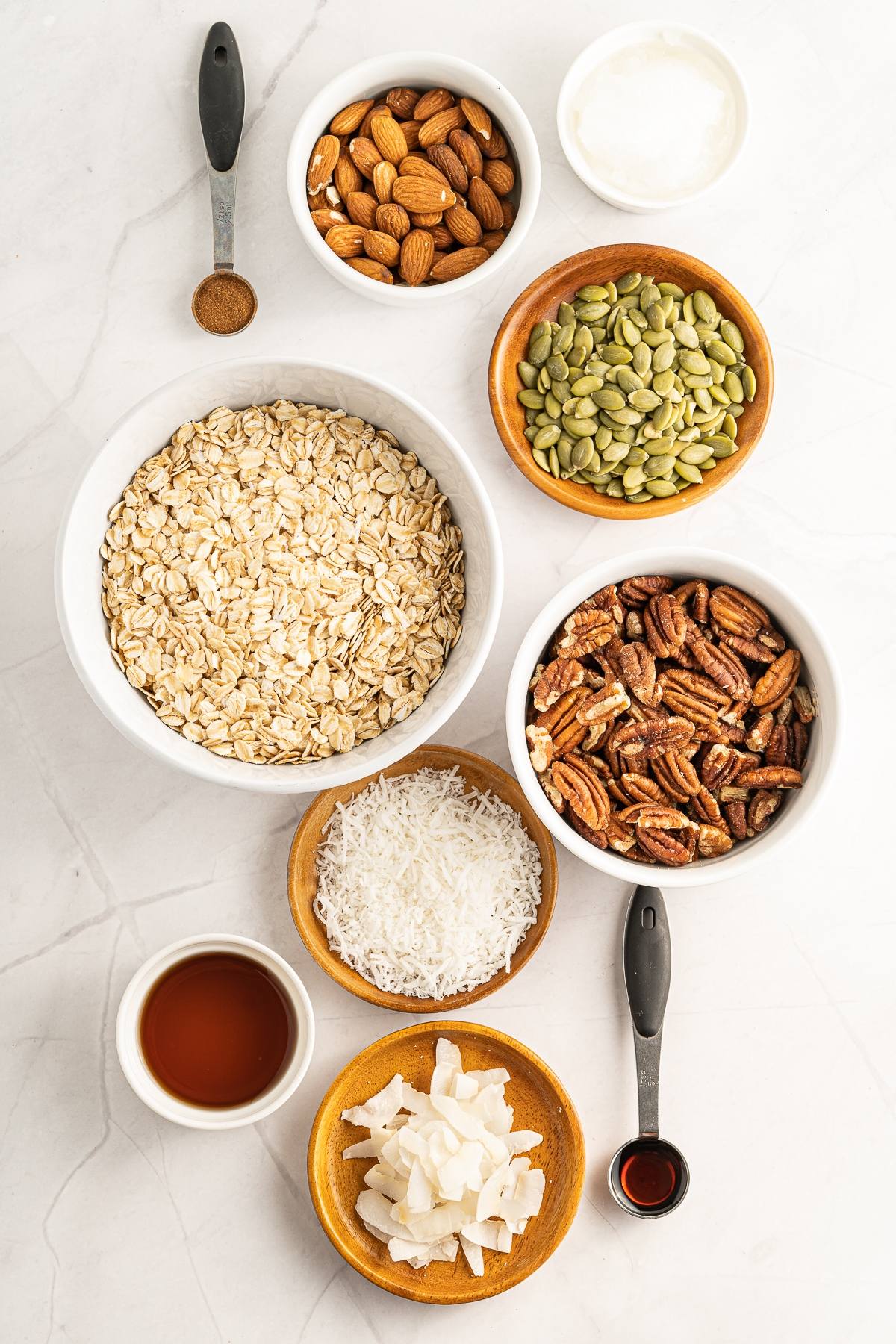 Assorted granola ingredients in bowls—oats, nuts, seeds, coconut, syrup, vanilla, and spices—on a marble surface, ready for homemade granola that's part of a healthy breakfast.