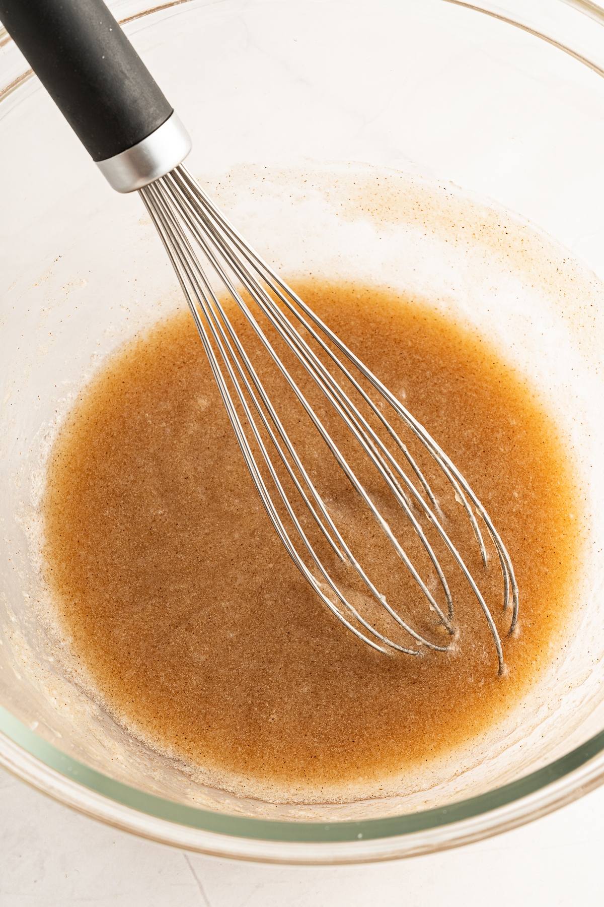 A whisk in a glass bowl mixing a light brown liquid batter on a white surface, perfect for easy homemade granola.