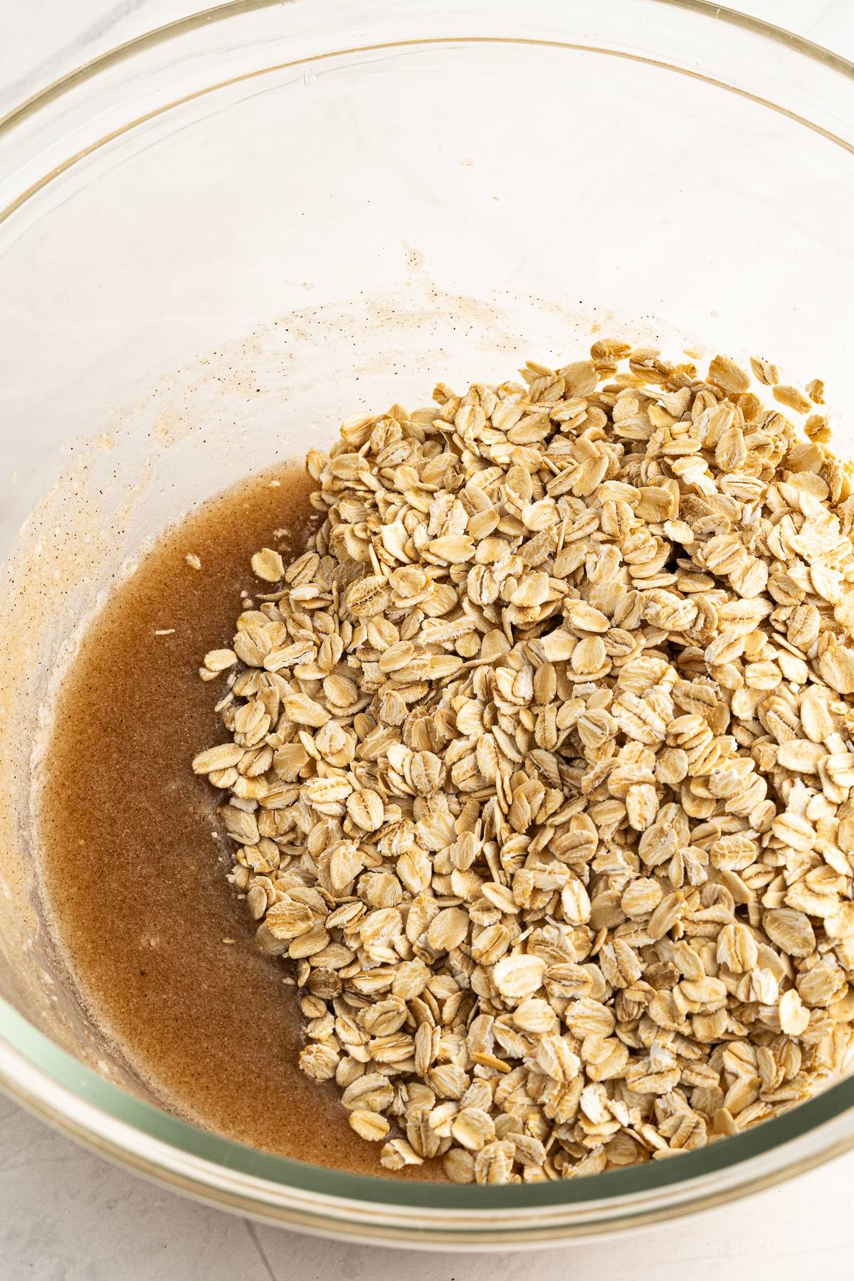 Rolled oats mixed with a brown liquid in a clear glass bowl on a light surface, perfect for making homemade granola and part of a healthy breakfast.