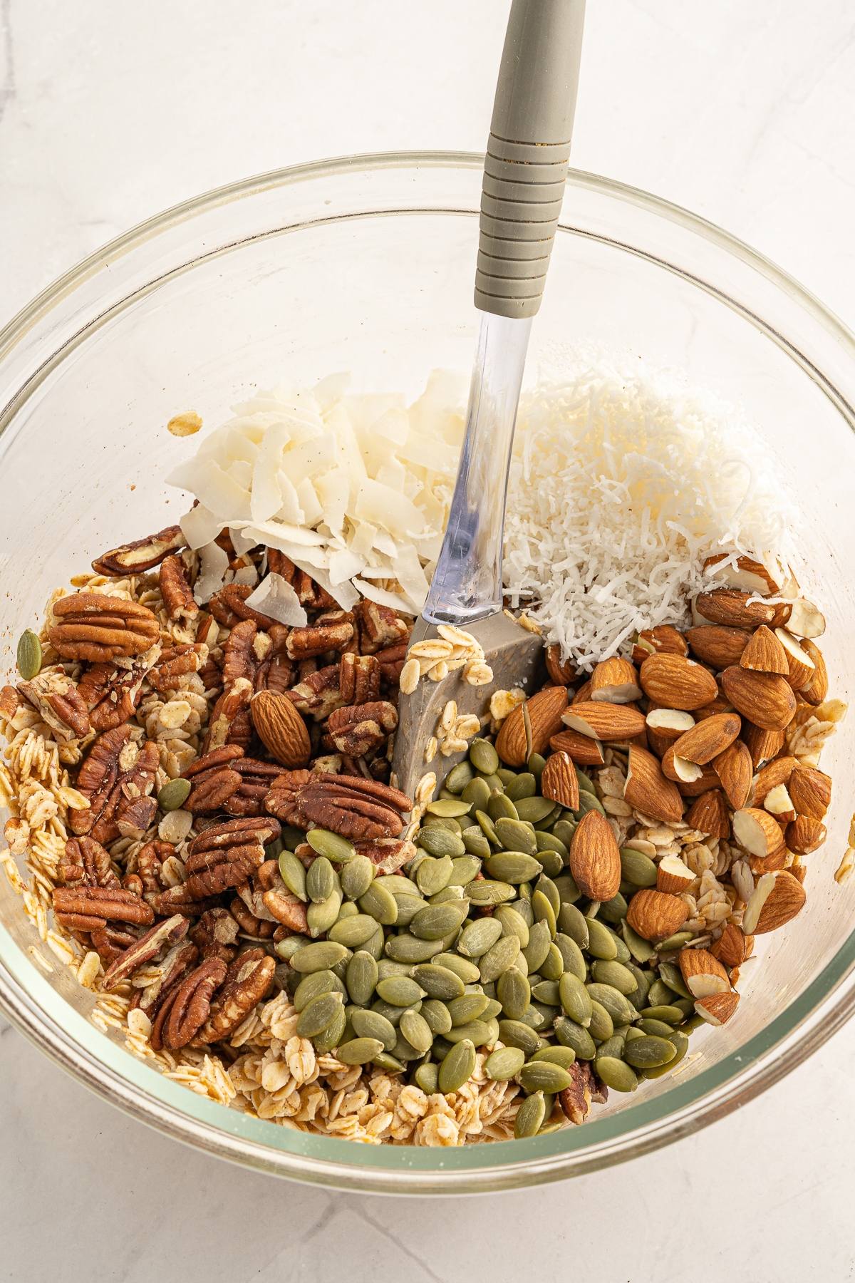 A glass bowl with oats, pecans, pumpkin seeds, almonds, coconut flakes, and shredded coconut being mixed—perfect for an easy homemade granola that’s part of a healthy breakfast.