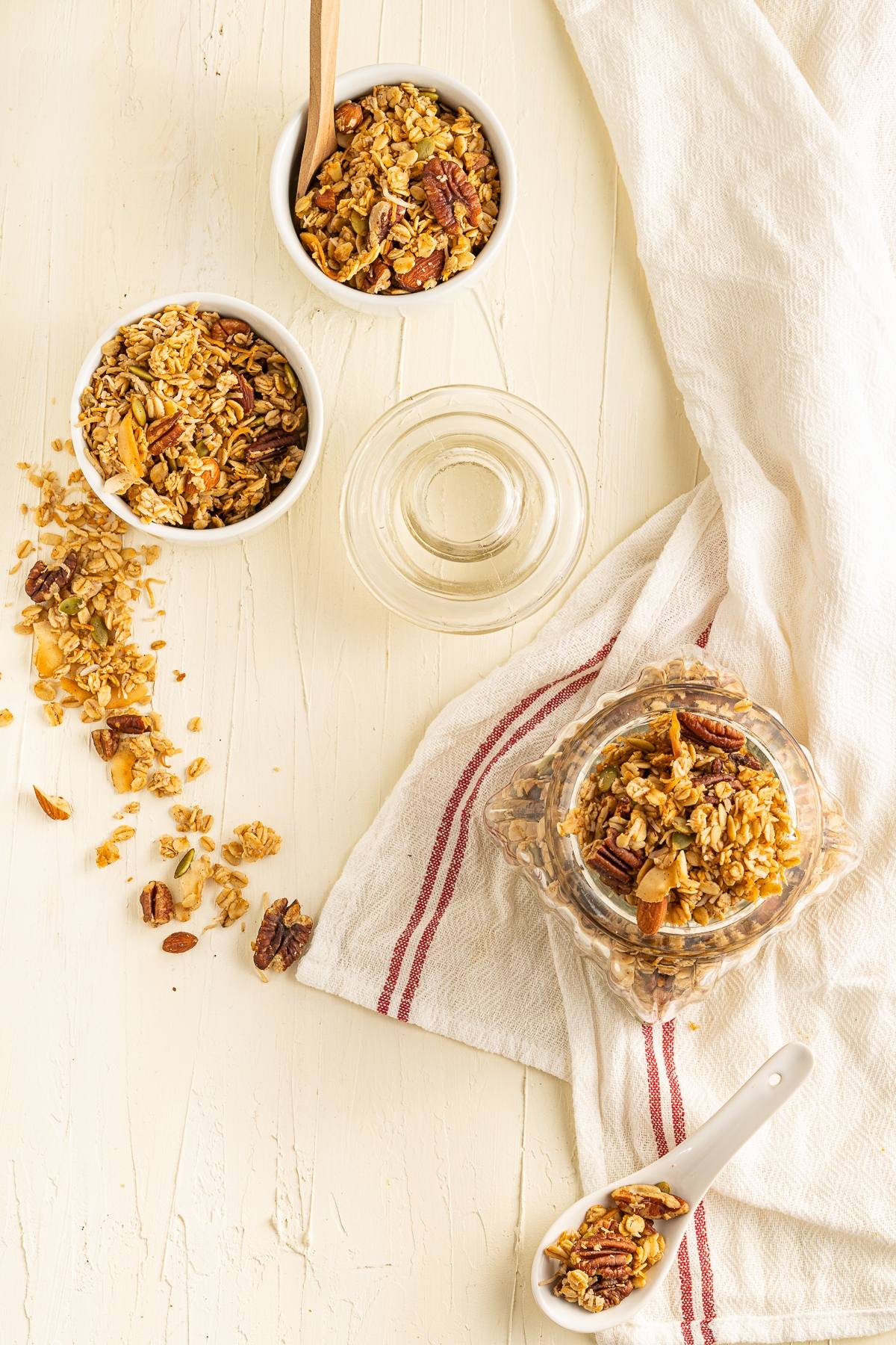 Jars and bowls of homemade granola with a spoon on a white table, next to a white cloth with a red stripe—perfect as part of a healthy breakfast.